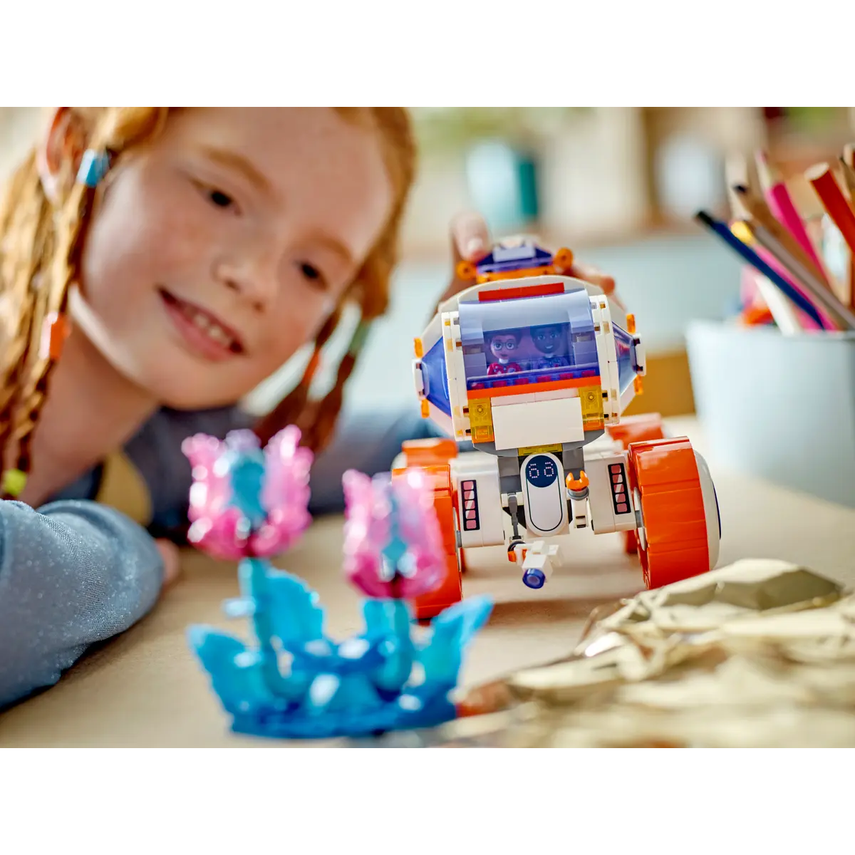 Una ragazza dai capelli rossi sorride, guardando un rover Lego con due personaggi all'interno. Fiori Lego rosa e blu in primo piano.
