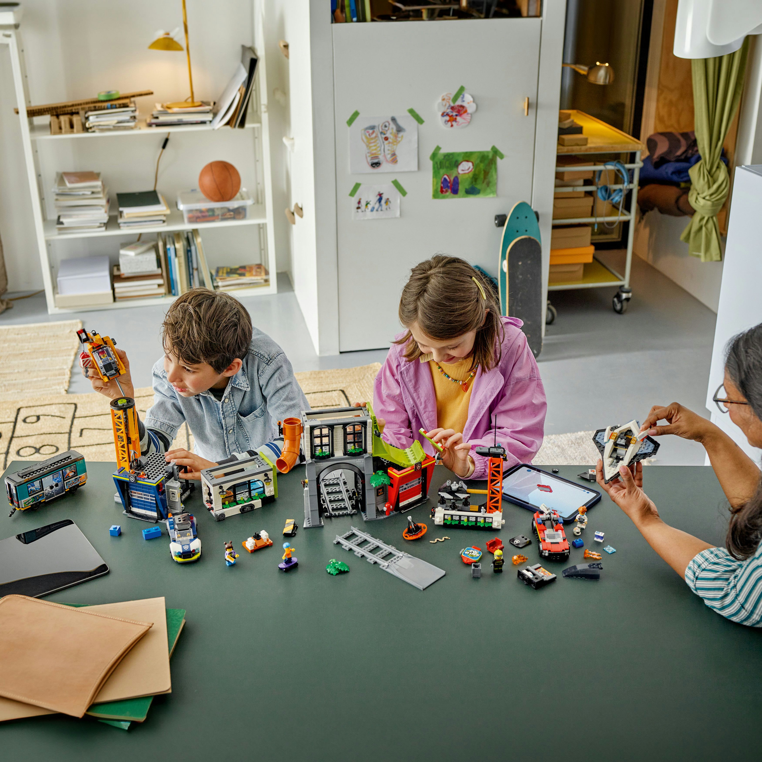 Üç çocuk ve bir yetişkin yeşil bir masada Lego setleri topluyor. Oyuncaklar ve tabletler masaya dağılmış durumda.