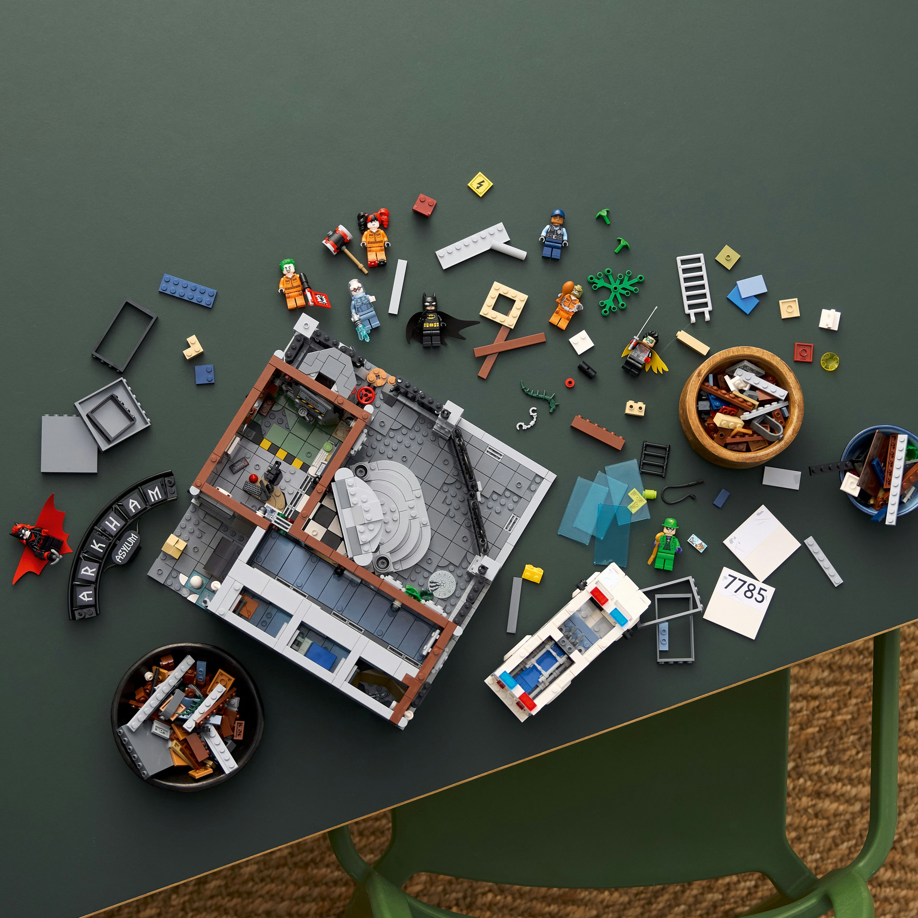 Zestaw Lego na zielonym stole, z postaciami Batmana i klockami Arkham Asylum, z zielonym krzesłem.