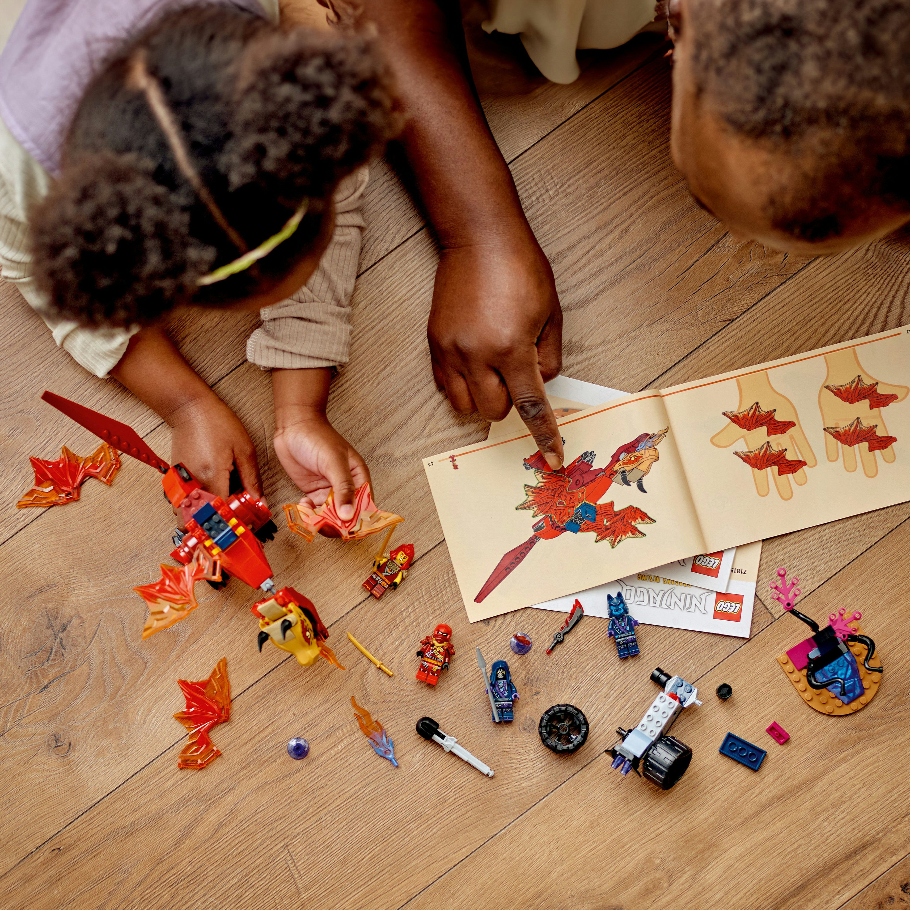 Dwie osoby budują zestaw Lego. Smok, figurki i instrukcje są na drewnianej podłodze.