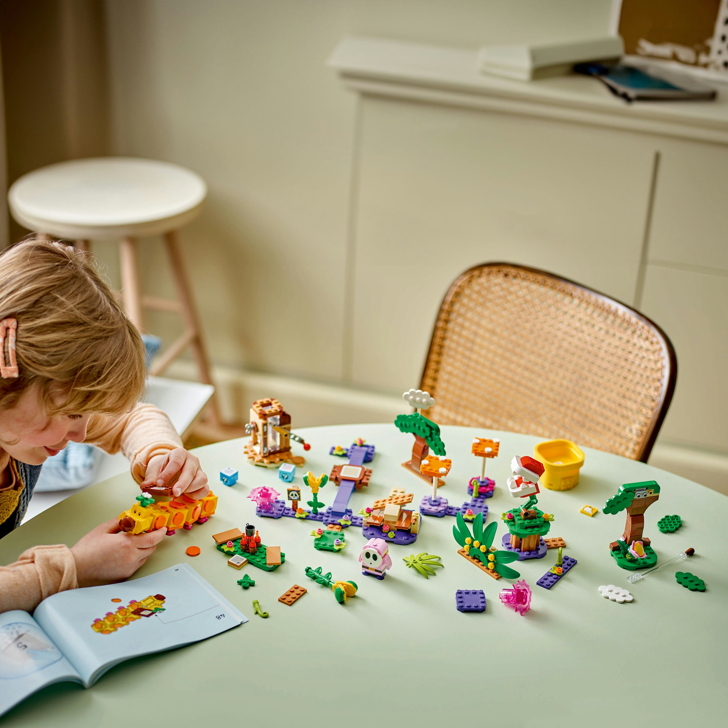 Une fille assemble un jeu Lego sur une table verte. L'ensemble est une scène colorée avec des arbres et des figurines.
