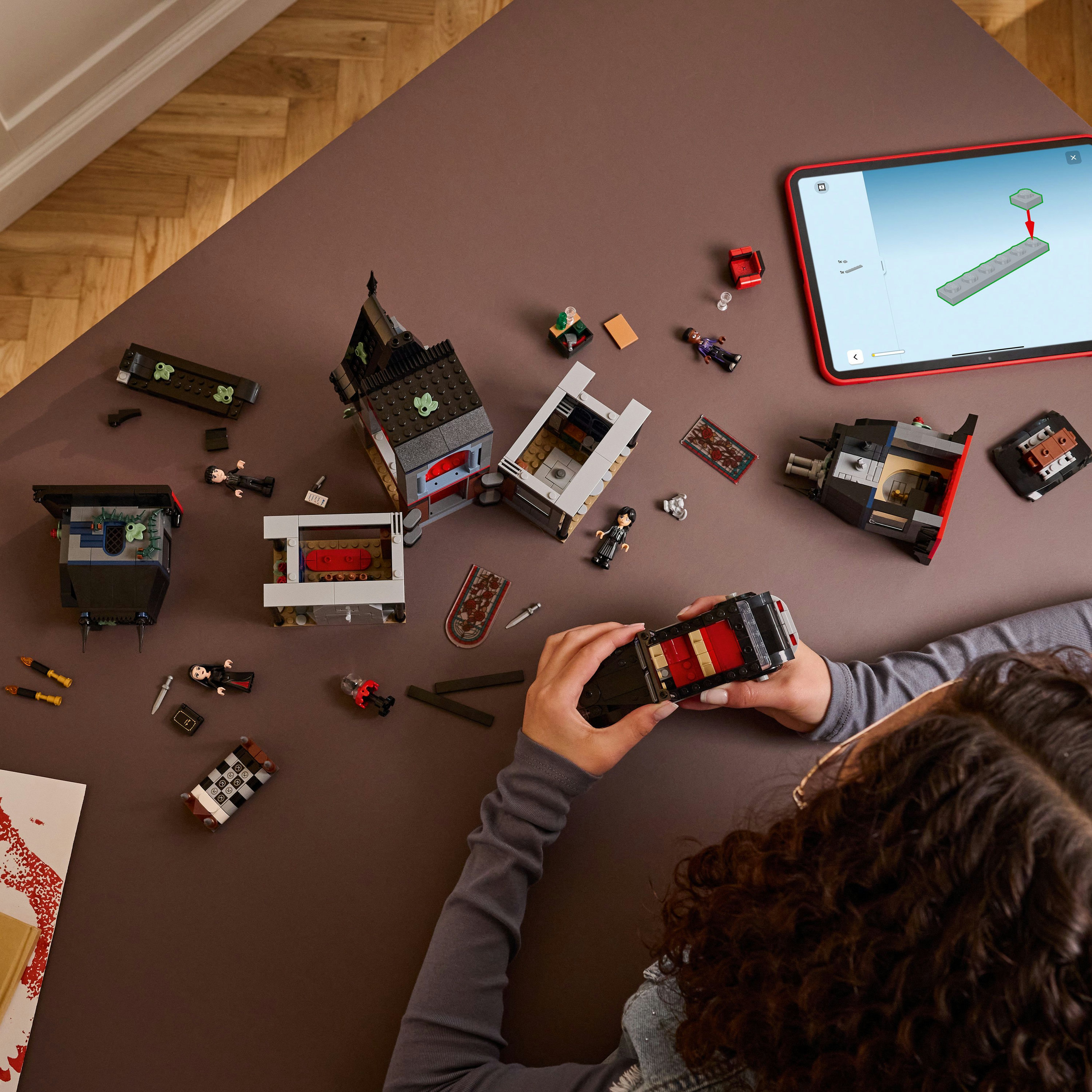 Osoba składa zestaw Lego, z instrukcjami na tablecie. Różne klocki Lego i minifigurki są rozrzucone.