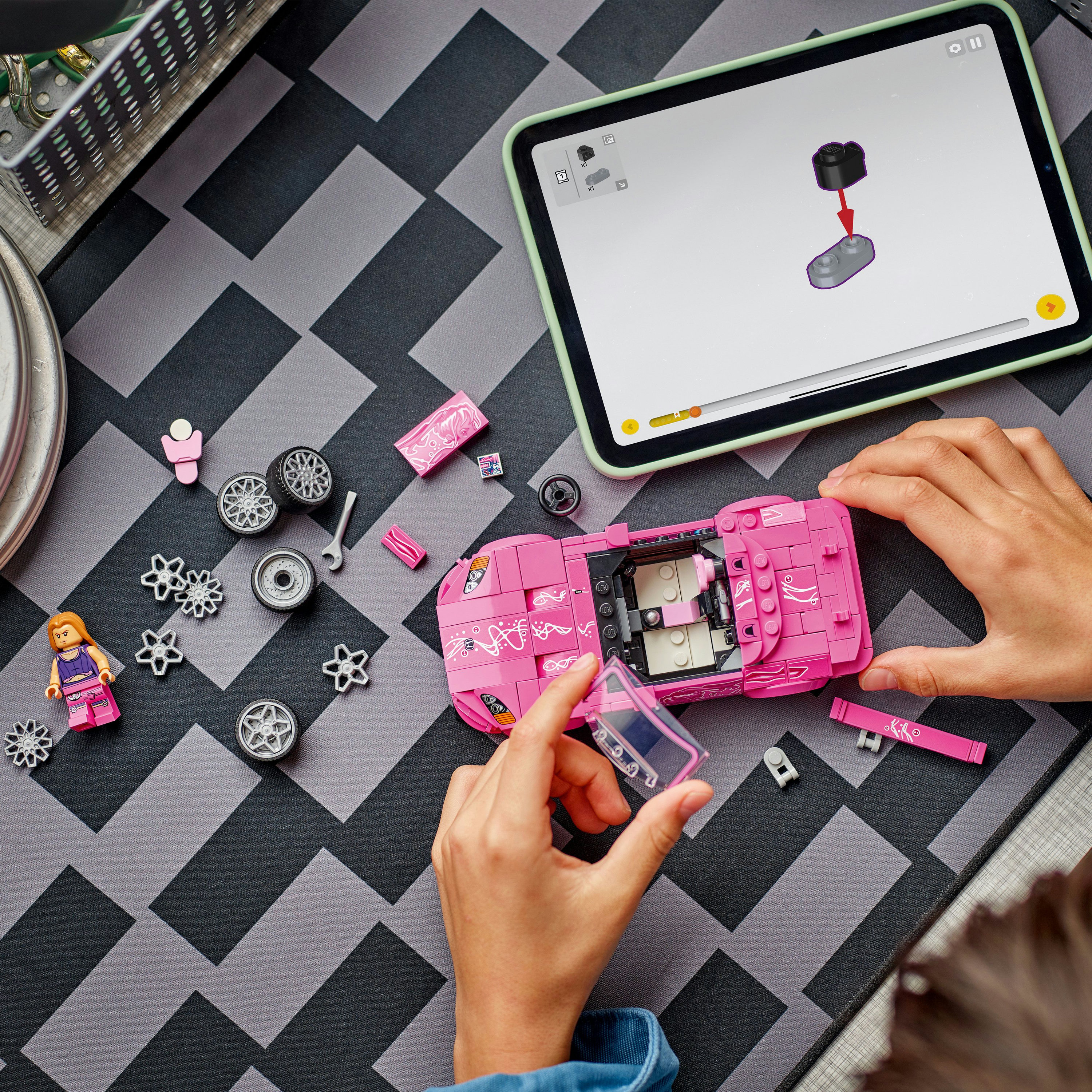 Osoba buduje różowy samochód Lego. Części i instrukcje znajdują się na stole, w tym tablet z cyfrowymi instrukcjami.