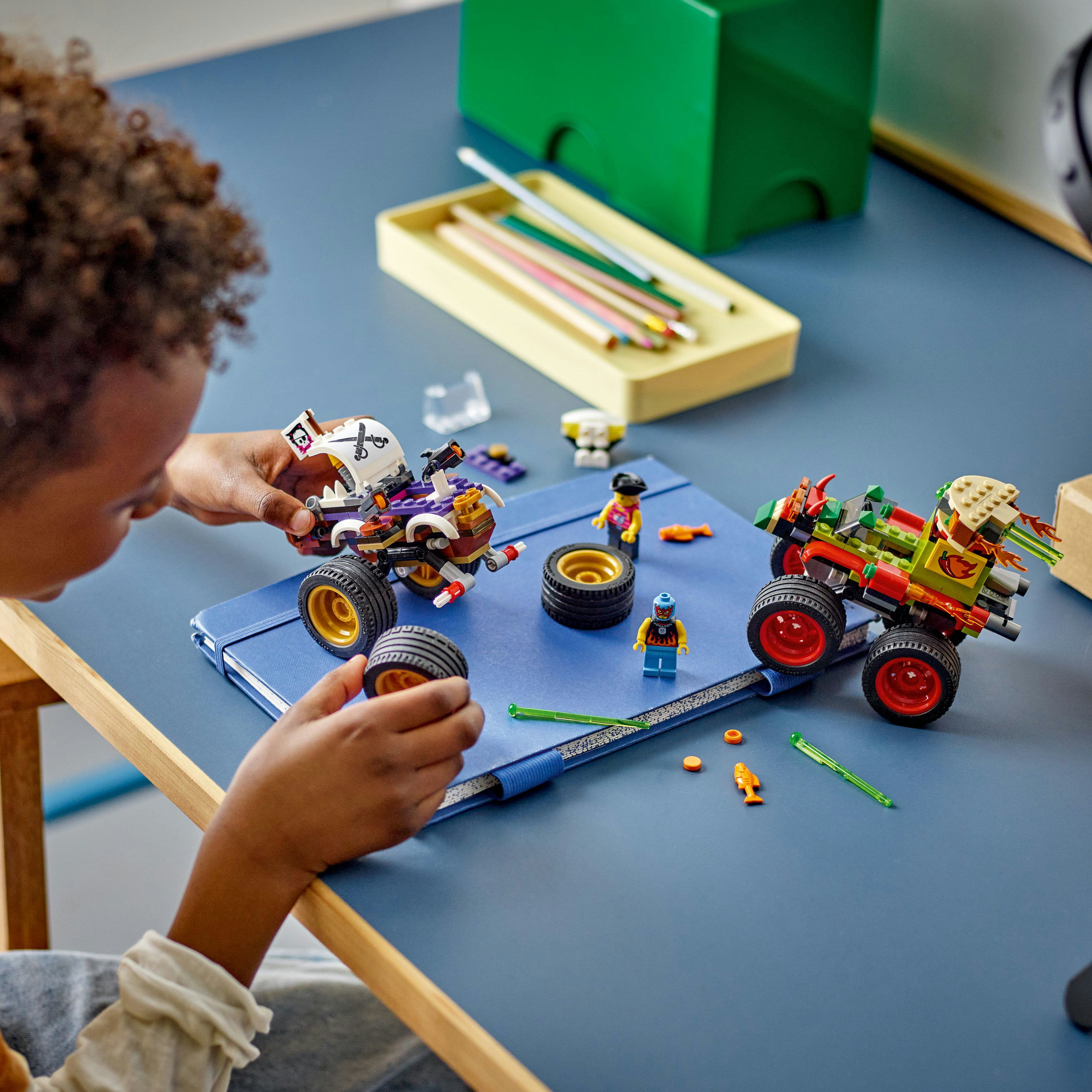 Un enfant joue avec des monster trucks Lego sur un bureau bleu. Des figurines et pièces Lego sont éparpillées.