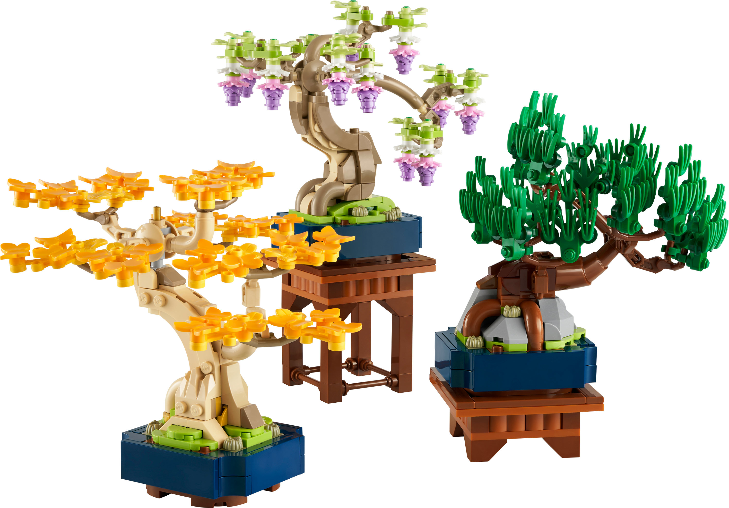 Drie Lego bonsai bomen in verschillende kleuren op blauwe bases en bruine stands. Eén heeft gele bladeren, een ander roze bloemen.