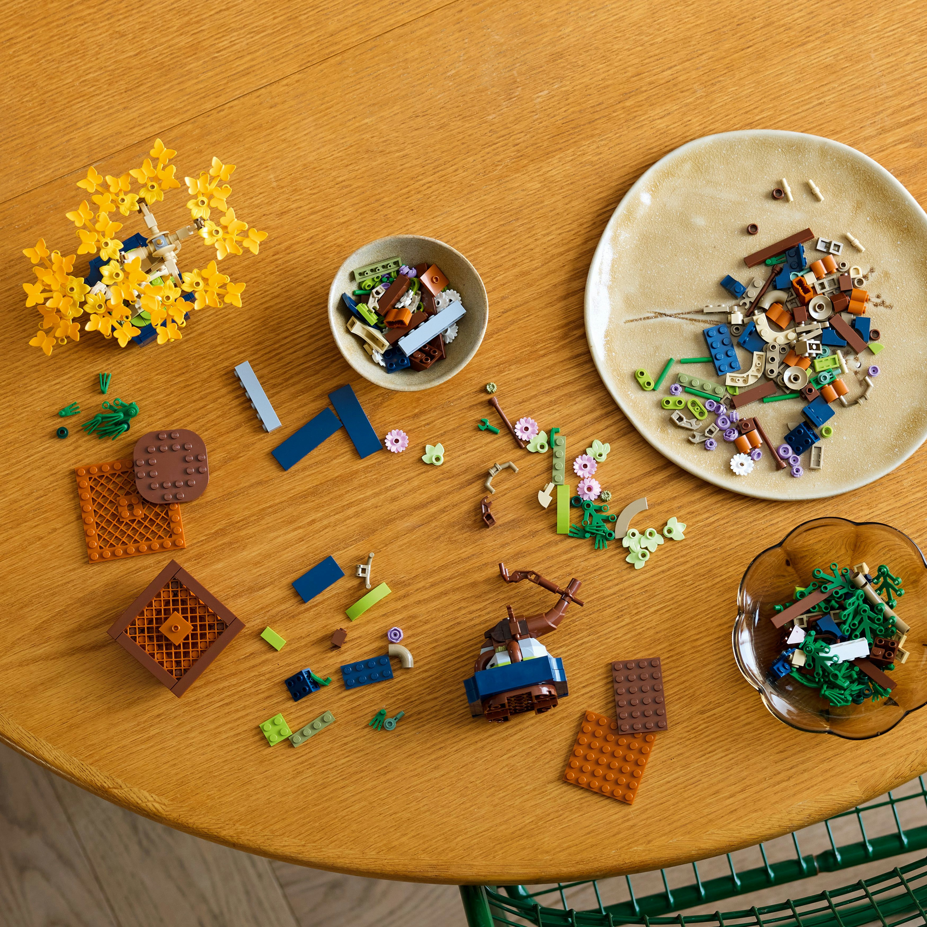 Houten tafel met verspreide Lego-stukken, kommen en een bord. Inclusief gele bloemen en een groene stoel.