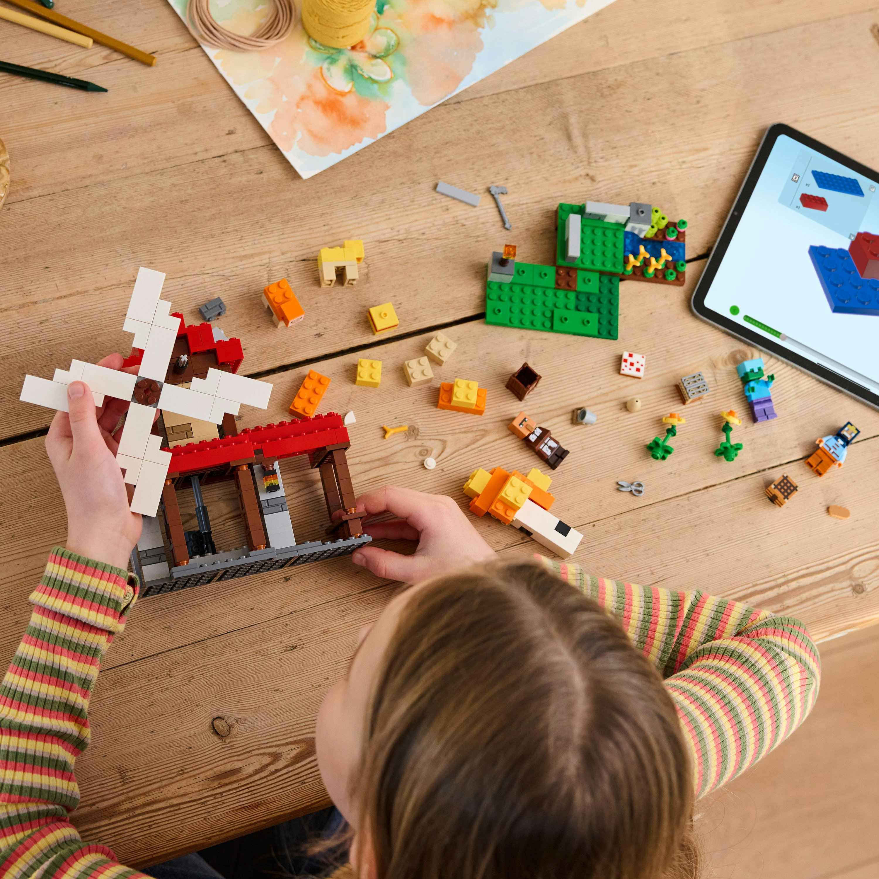 Dziecko składa wiatrak Lego. Inne klocki Lego i tablet z instrukcjami leżą na drewnianym stole.