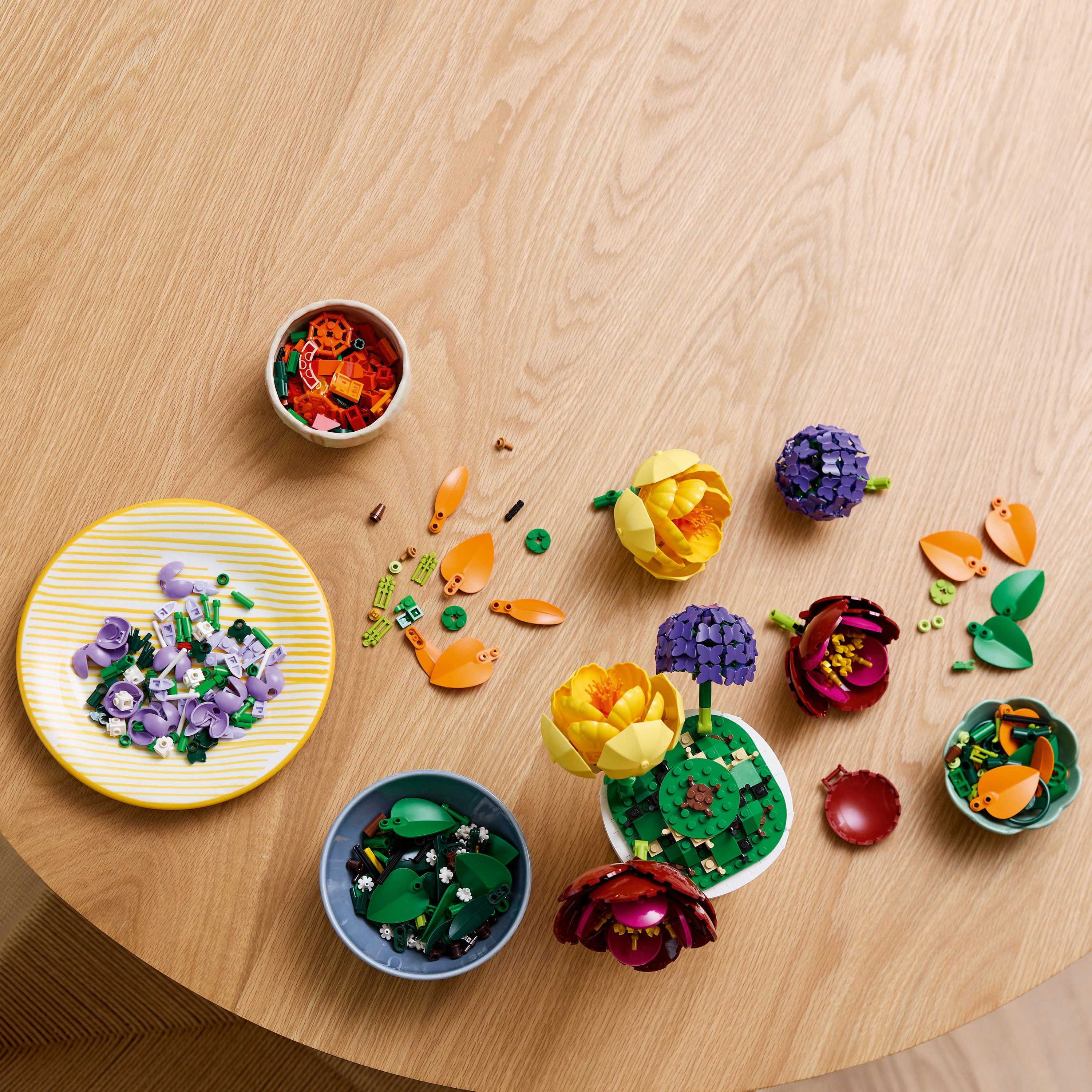 Table en bois avec des fleurs Lego et des parties de fleurs dans des bols et une assiette. Les fleurs sont jaunes, violettes et rouges.