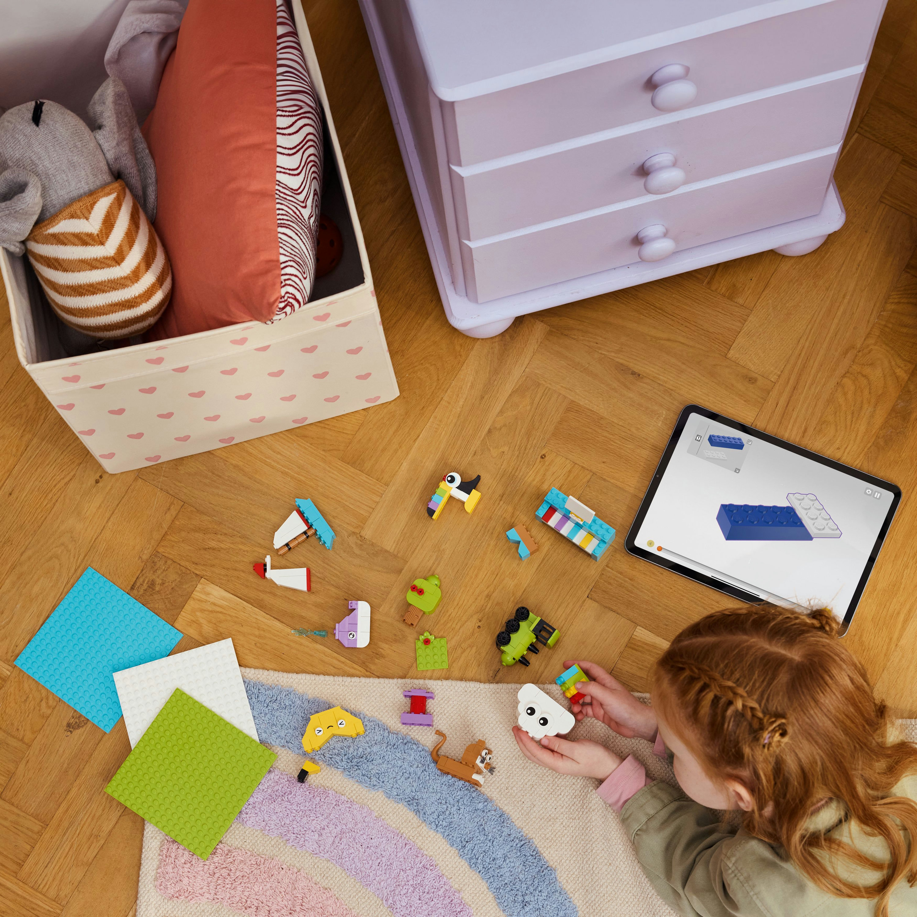 Dziecko bawi się klockami Lego na dywanie. Tablet wyświetla instrukcje. Szuflada i pudełko są w tle.