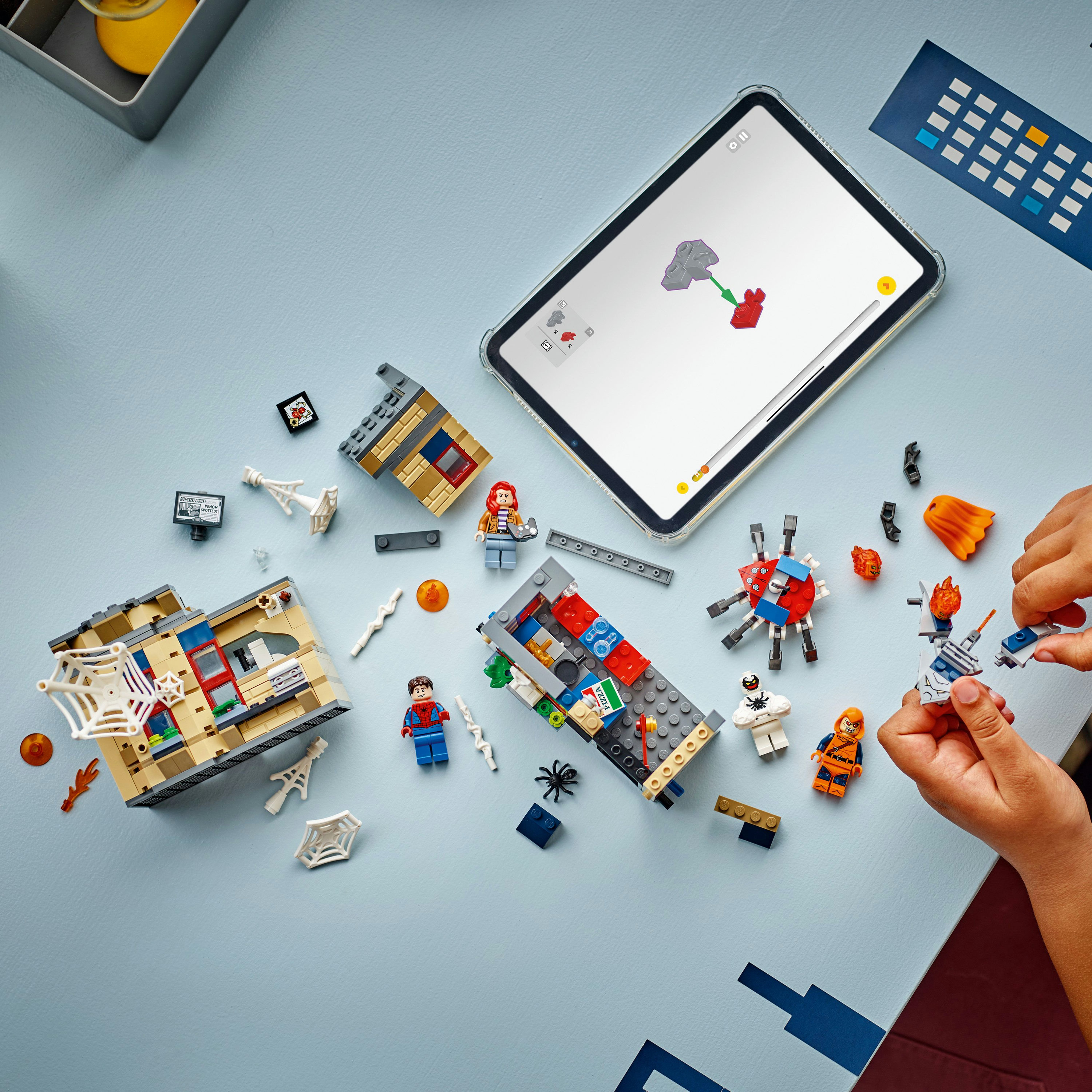 Zestaw Lego jest montowany na niebieskiej powierzchni. Widoczne są figurki Lego, budynki i iPad z instrukcjami.