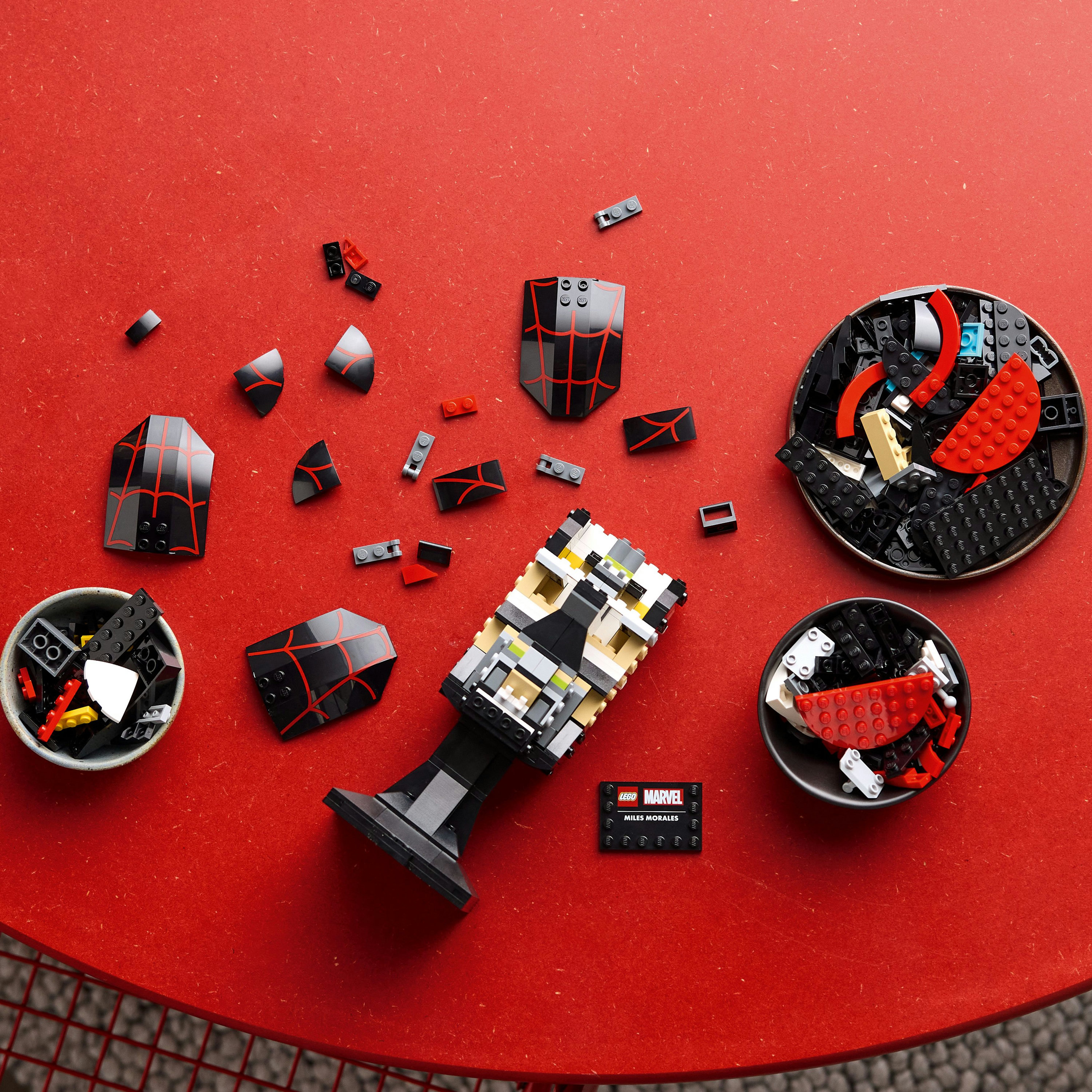 Ensemble Lego sur table rouge, comprend pièces de masque Spider-Man, briques Lego dans bols et enseigne Lego Marvel.