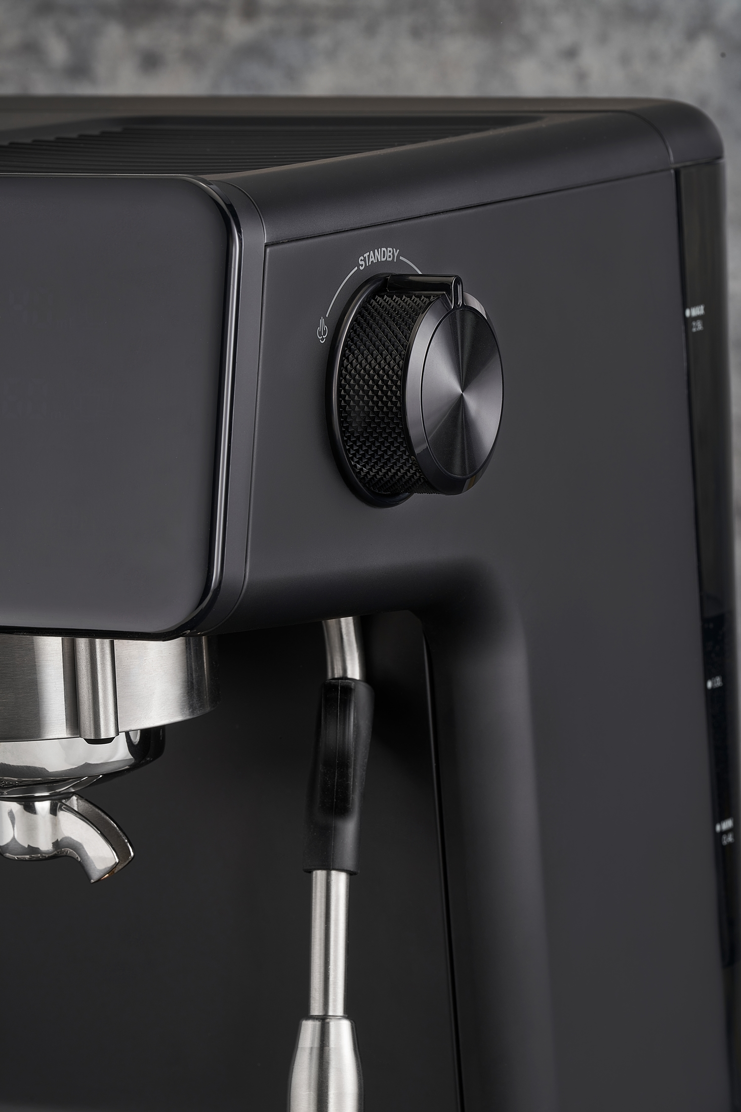 Nahaufnahme einer schwarzen Espressomaschine. Ein Drehknopf zeigt "Standby". Ein Siebträger und eine Dampflanze sind sichtbar. Der Hintergrund ist eine graue Wand.