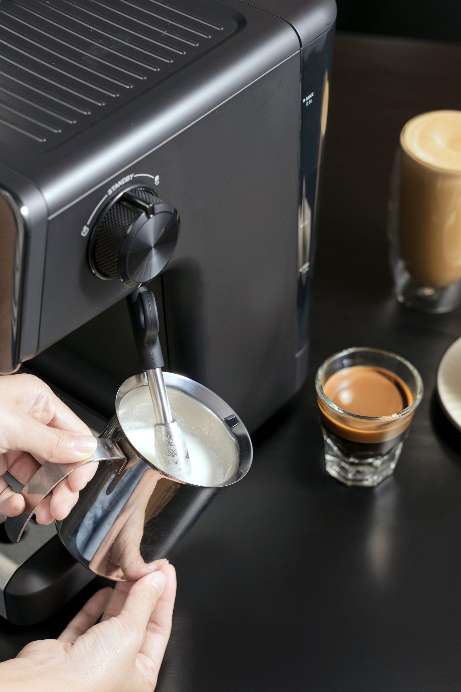 Eine Person schäumt Milch mit einer Espressomaschine auf. Espresso- und Latte-Gläser stehen auf dem Tisch.