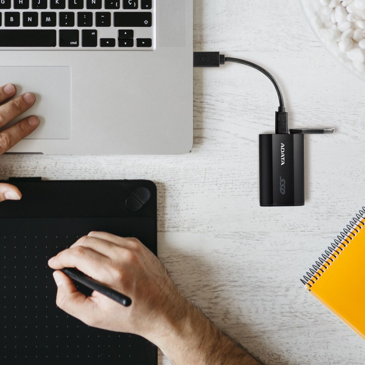 Laptop jest podłączony do zewnętrznego dysku twardego. Widoczne są dłonie osoby, jedna używająca tabletu, druga w pobliżu laptopa.