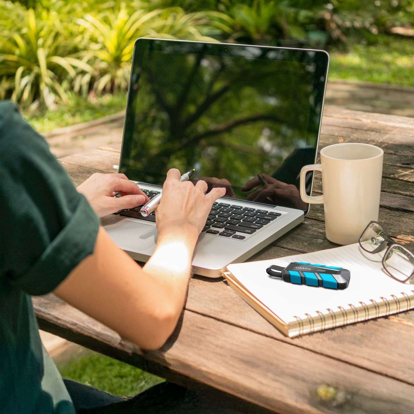 Personne tapant sur un ordinateur portable en extérieur, avec une tasse à café, des lunettes, un carnet et un SSD bleu et noir sur une table en bois.