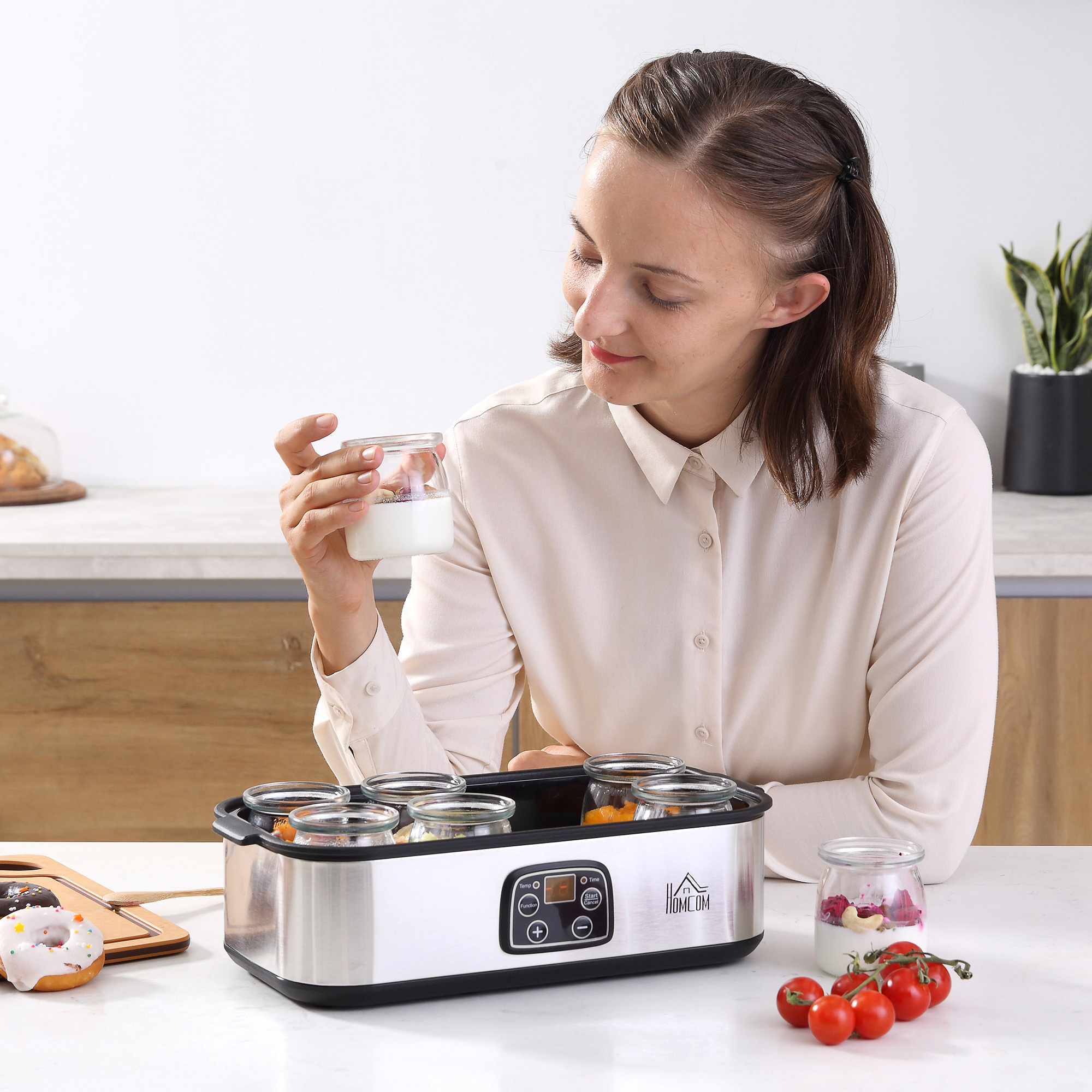 Una mujer mira un frasco de yogur. Hay una yogurtera, rosquillas y tomates en la encimera.