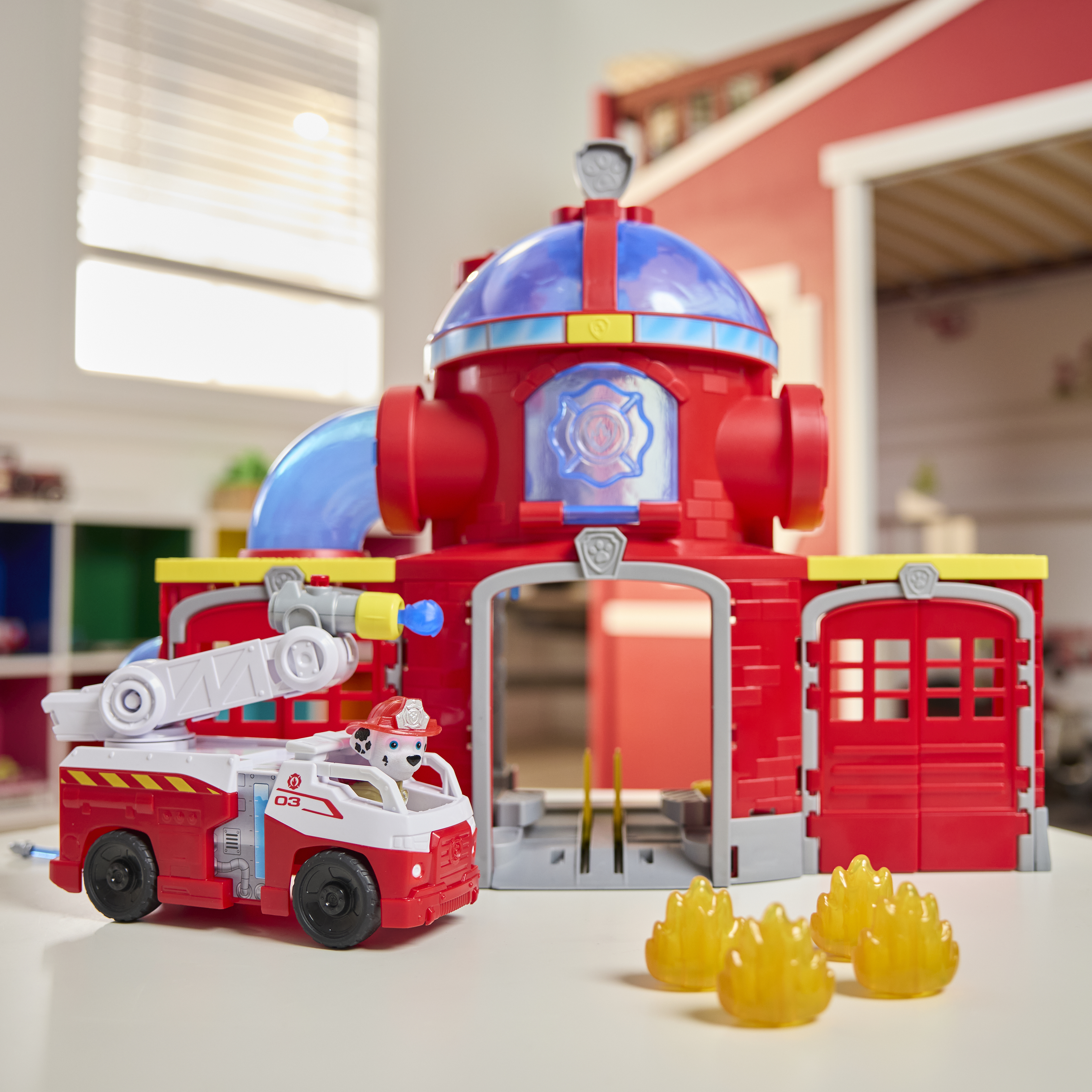 Czerwony zestaw zabawek z wozem strażackim i remizą Paw Patrol. W ciężarówce jest Marshall. Żółte zabawki w kształcie płomieni leżą na stole.
