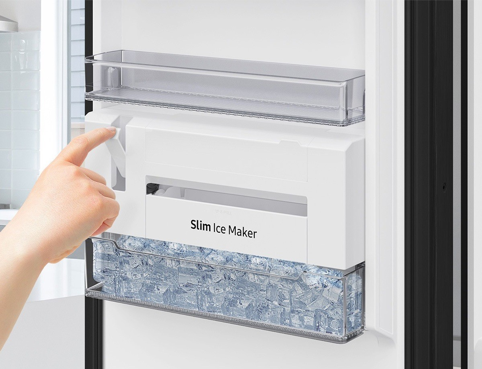 Una mano presiona una palanca en una máquina de hielo blanca dentro de un refrigerador. La máquina de hielo dice 'Slim Ice Maker' y contiene cubitos de hielo.