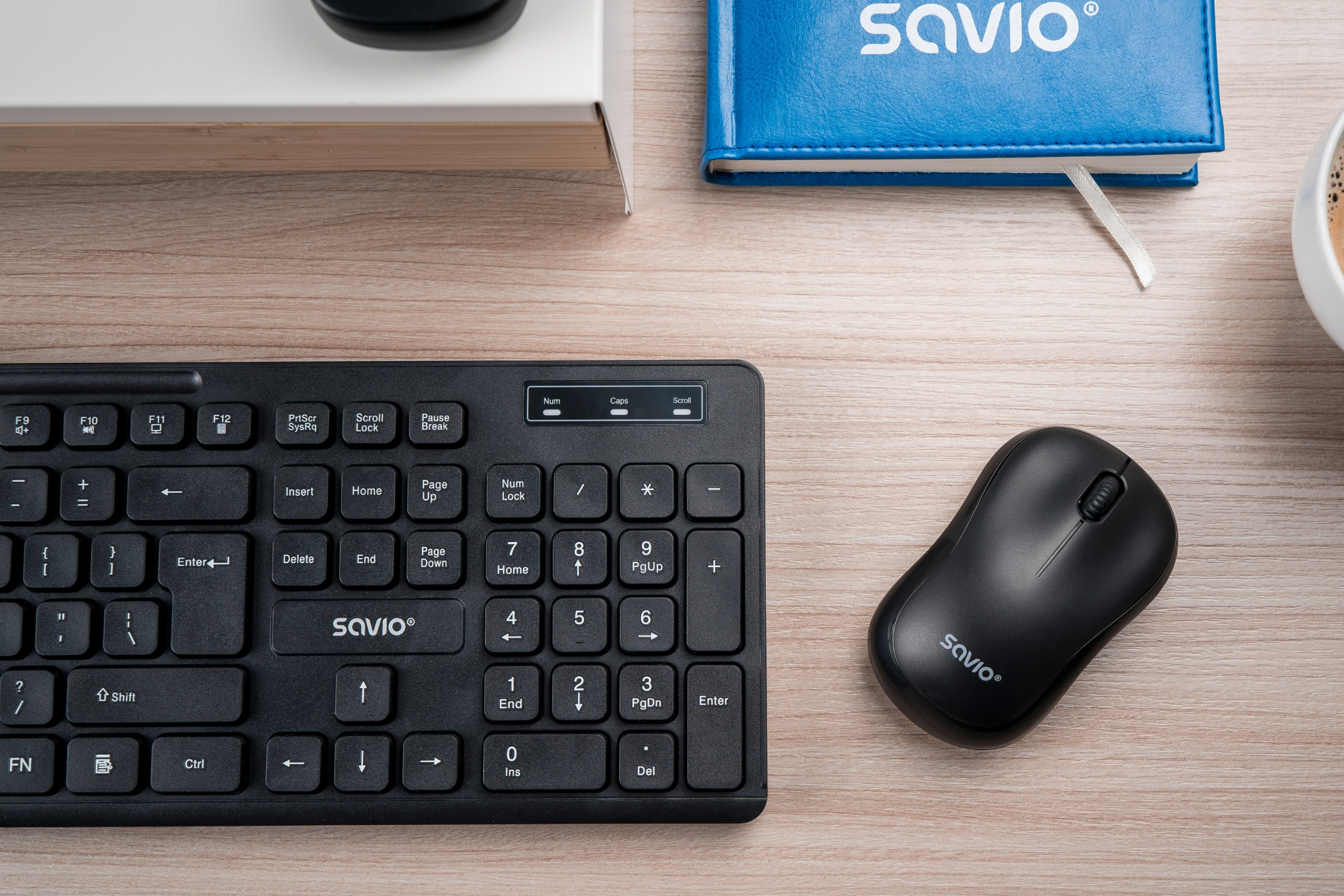 Czarna klawiatura, mysz i niebieski notatnik na drewnianym stole. Klawiatura ma markę SAVIO. Obecny jest również kubek kawy.