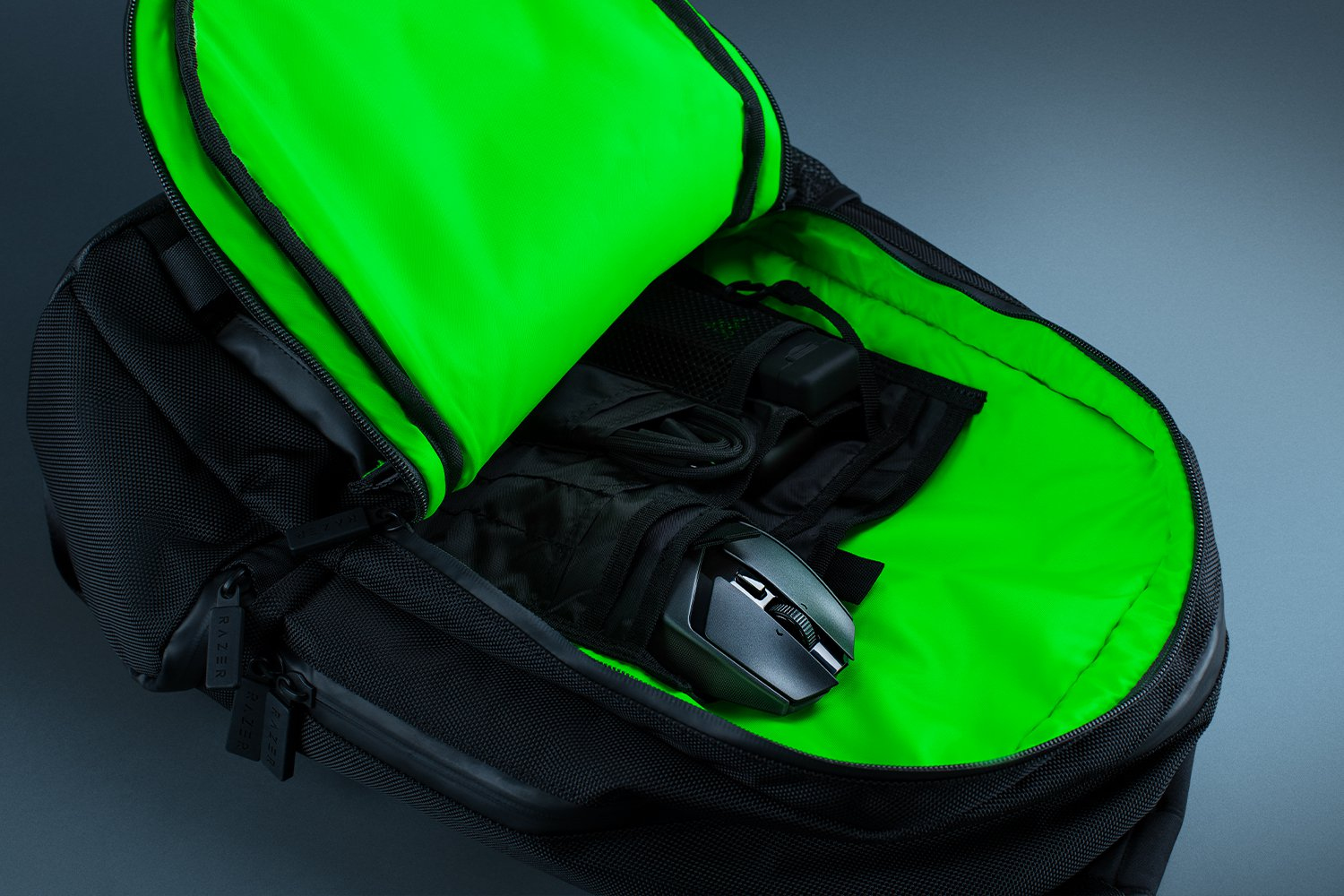 Czarny plecak Razer otwarty, ukazujący zielone wnętrze. Czarna mysz i inne akcesoria w środku.