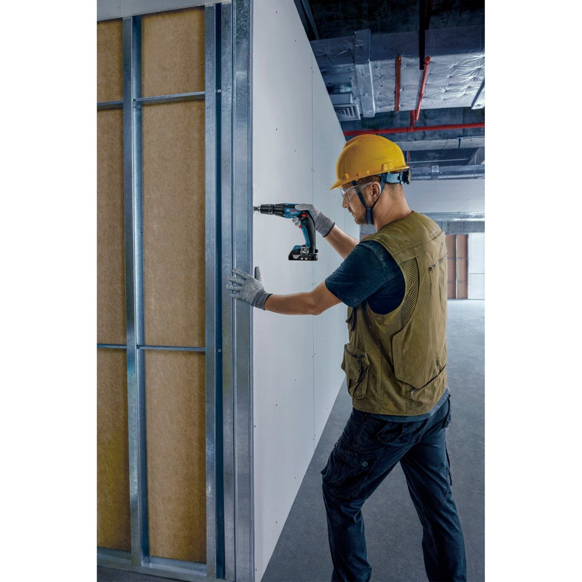 Robotnik budowlany wierci w ramie ściany, nosząc żółty kask, okulary ochronne i kamizelkę. Używa niebiesko-czarnej wiertarki.