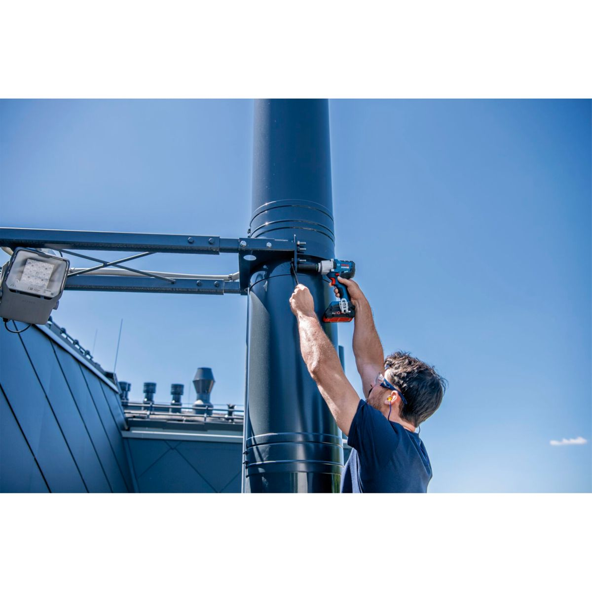 Mężczyzna w niebieskiej koszuli używa wiertarki na czarnym słupie z oprawą oświetleniową, na tle błękitnego nieba.