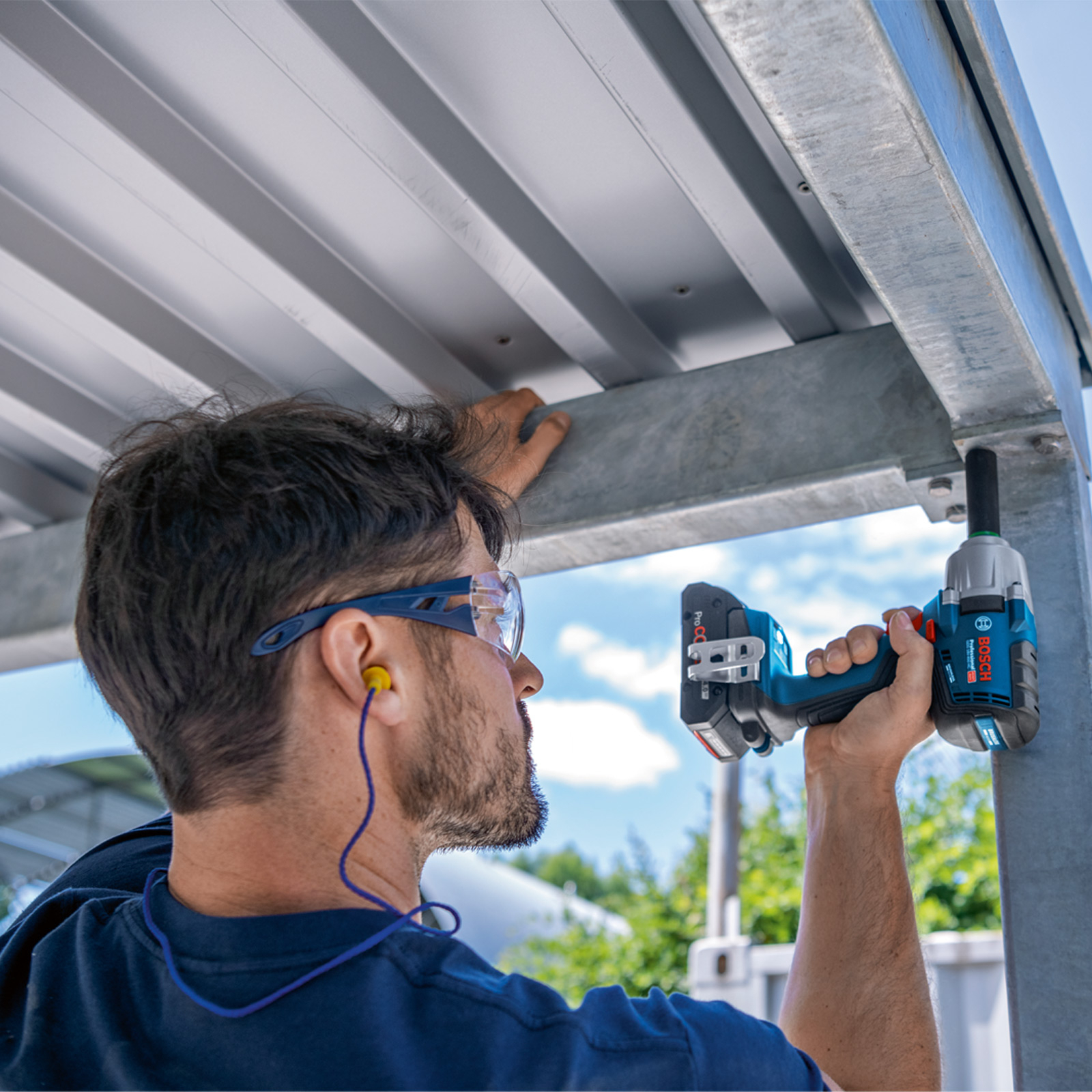 Mężczyzna używa niebiesko-czarnego elektronarzędzia do pracy przy metalowej konstrukcji. Nosi okulary ochronne i ochronniki słuchu.