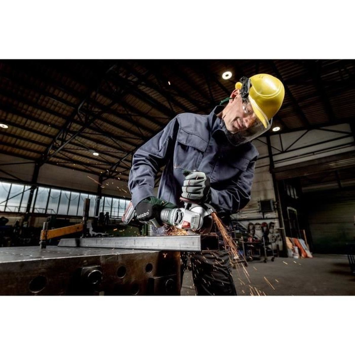 Robotnik szlifuje metal elektronarzędziem, lecą iskry. Ma na sobie żółty kask, okulary ochronne i rękawice w warsztacie.