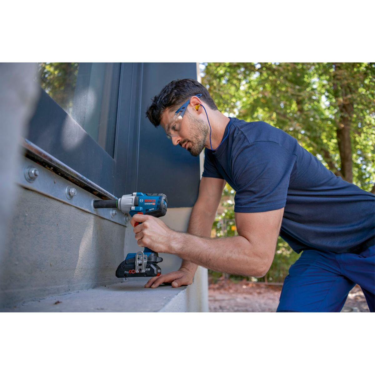 Mężczyzna w niebieskiej koszuli używa wiertarki. Ma na sobie okulary ochronne i zatyczki do uszu. Metalowa konstrukcja w tle.