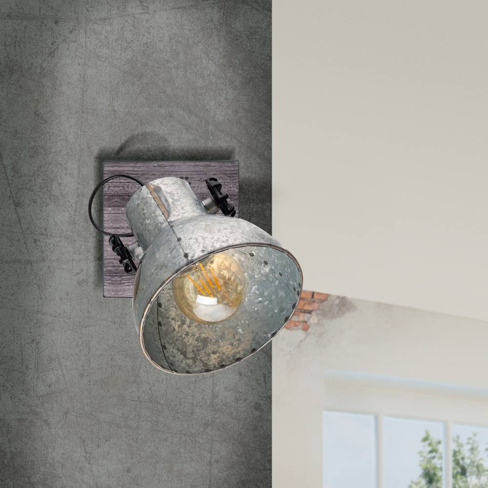 Eine industrielle Metalllampe ist auf einer Holzplatte an einer strukturierten grauen Wand montiert. Eine warme Glühbirne ist sichtbar.