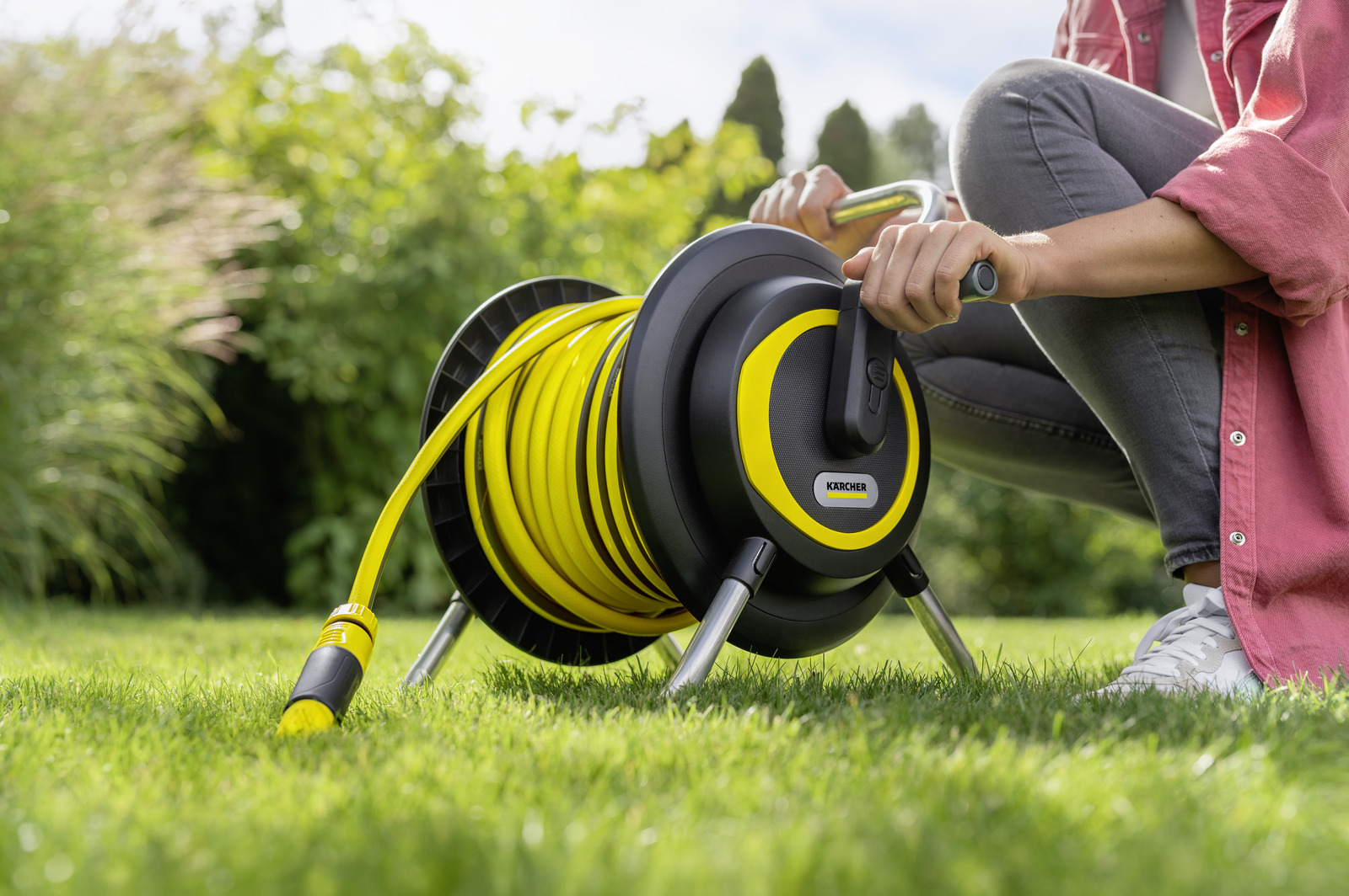 Osoba zwija żółty wąż na czarno-żółtej szpuli w trawiastym ogrodzie. Tło przedstawia zielone liście.