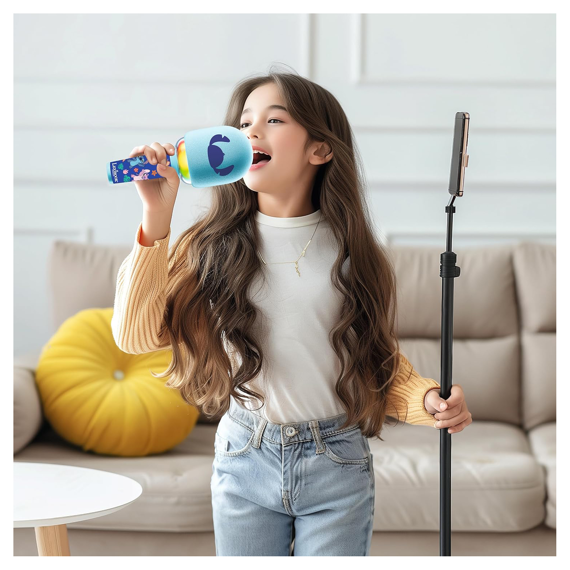 Dziewczyna śpiewa do mikrofonu. Ma na sobie dżinsy i sweter. Obok niej znajduje się stojak na telefon, a w tle sofa.