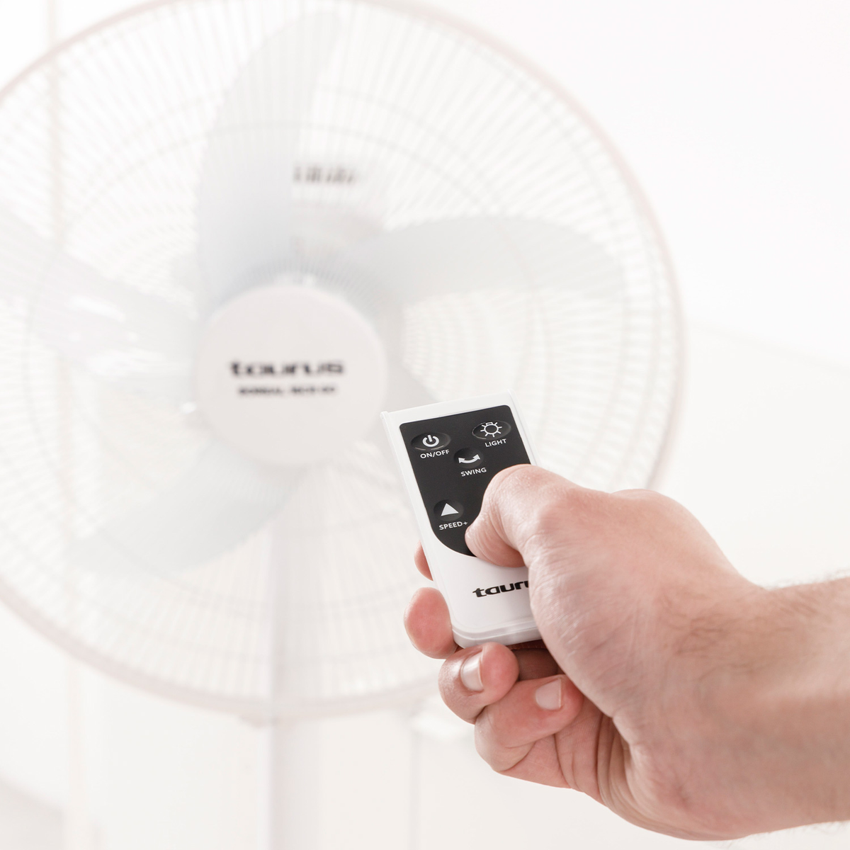 Ręka trzyma pilota w kierunku białego wentylatora. Pilot ma przyciski zasilania, światła, obrotu i prędkości. Wentylator jest w tle.