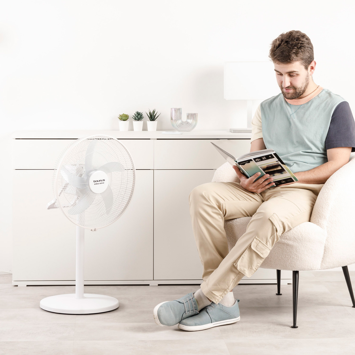 Mężczyzna siedzi na krześle i czyta książkę. Po lewej stronie znajduje się biały wentylator. Za nim biała szafka z roślinami.