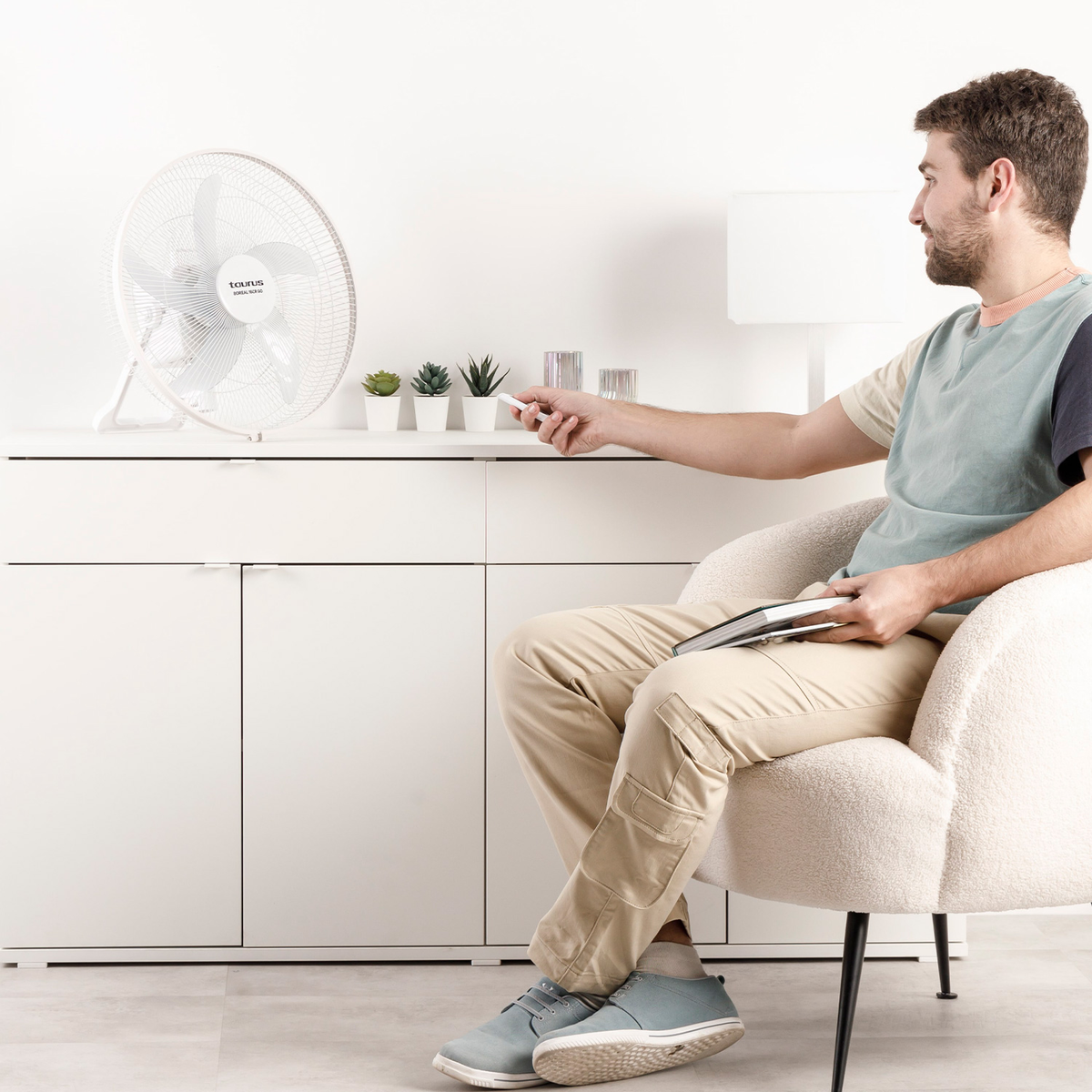 Mężczyzna siedzi, trzyma pilota, patrzy na wentylator. Biała szafka z roślinami. Biały wentylator po lewej. Beżowy fotel.