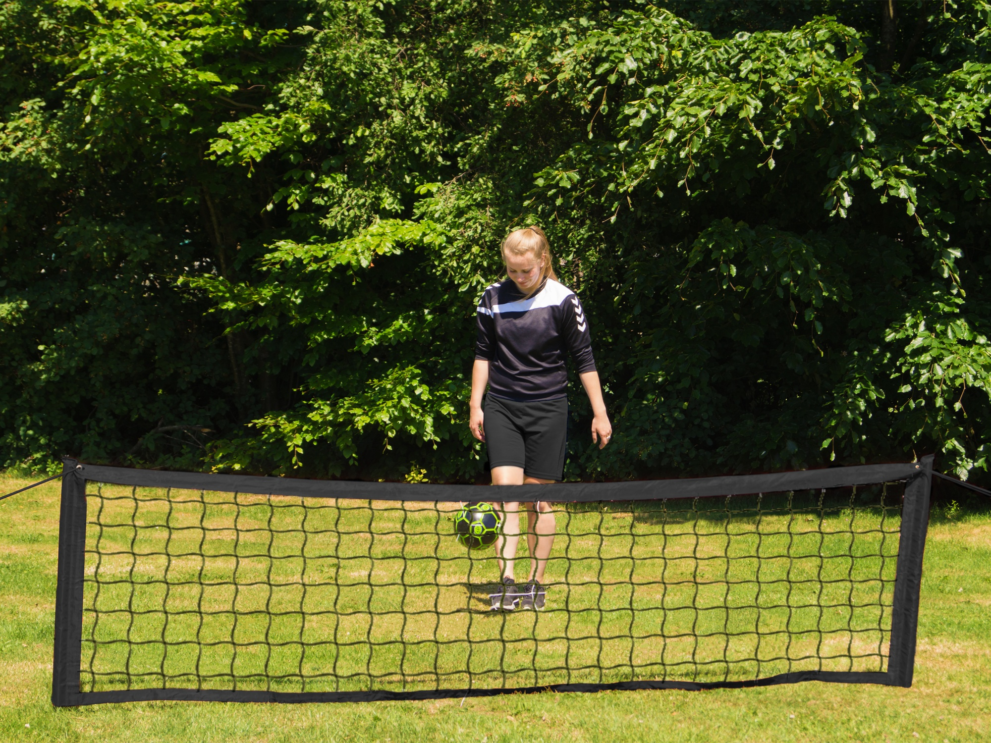 Ein Mädchen in Sportkleidung tritt gegen einen Fußball. Vorne ist ein Netz. Grünes Gras und Bäume sind im Hintergrund.
