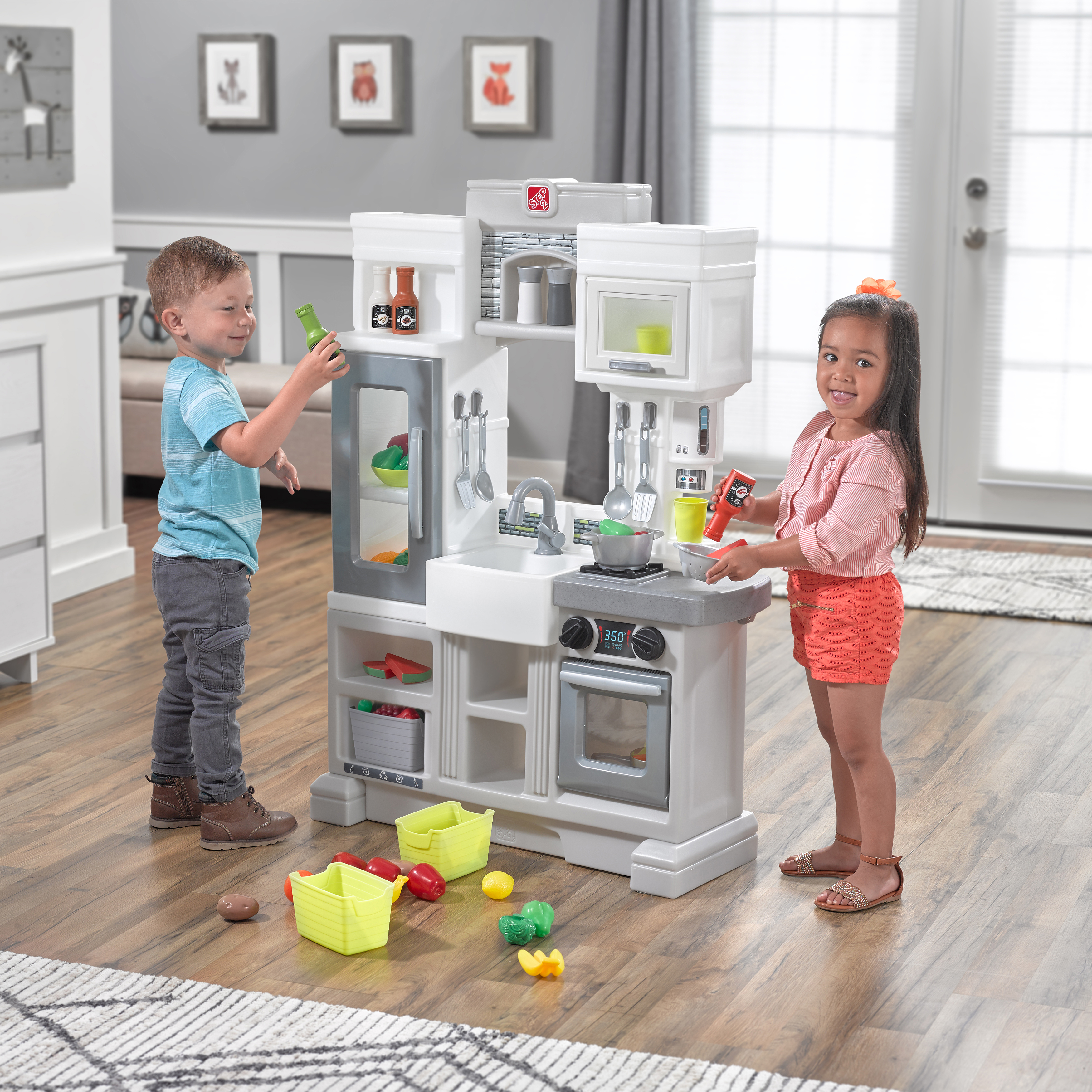 Zwei Kinder spielen mit einer Spielzeugküche und halten Spielzeugessen. Die Küche ist grau und weiß, auf einem Holzboden.