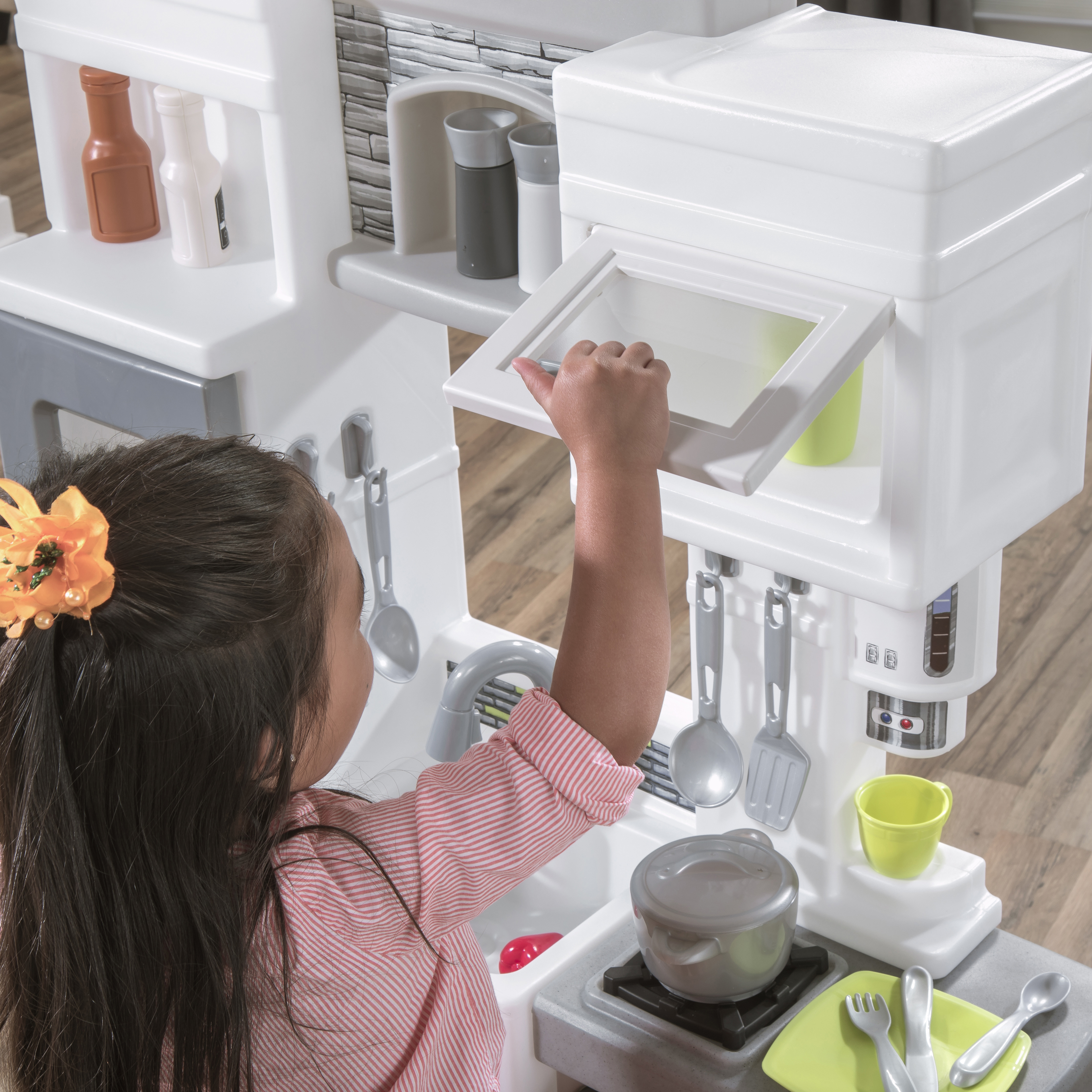 Ein Mädchen öffnet einen Spielzeugküchenschrank. Eine Spielzeugküche mit Utensilien und Lebensmitteln ist sichtbar. Ihr Haar hat eine Blume.