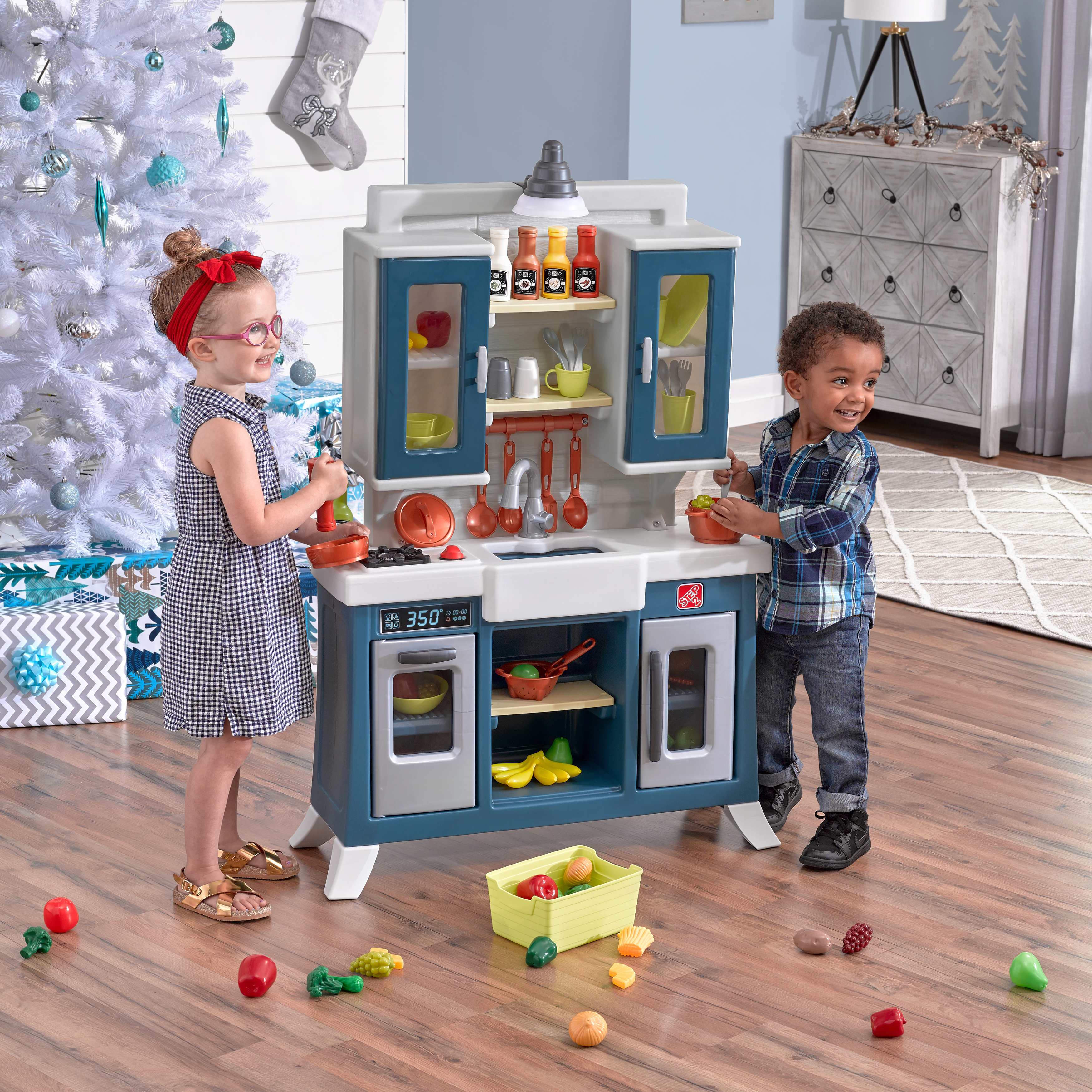 Zwei Kinder spielen mit einer Spielzeugküche. Im Hintergrund sind ein Weihnachtsbaum und Geschenke. Spielzeug liegt auf dem Boden verstreut.