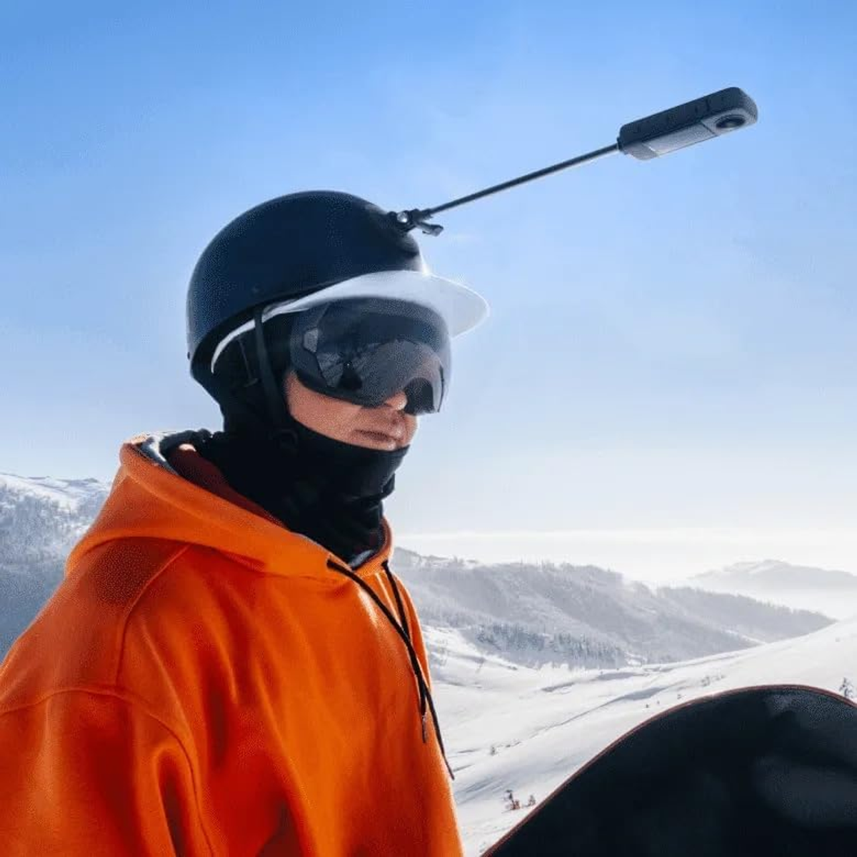 Osoba w pomarańczowej bluzie z kapturem i kasku z kamerą, goglami i snowboardem, stojąca na zaśnieżonej górze, z czystym, błękitnym niebem.