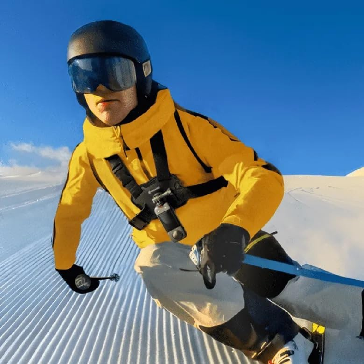 Narciarz w żółtej kurtce i czarnym kasku zjeżdża ze śnieżnego stoku. Niebieskie niebo w tle. Dołączona kamera GoPro.