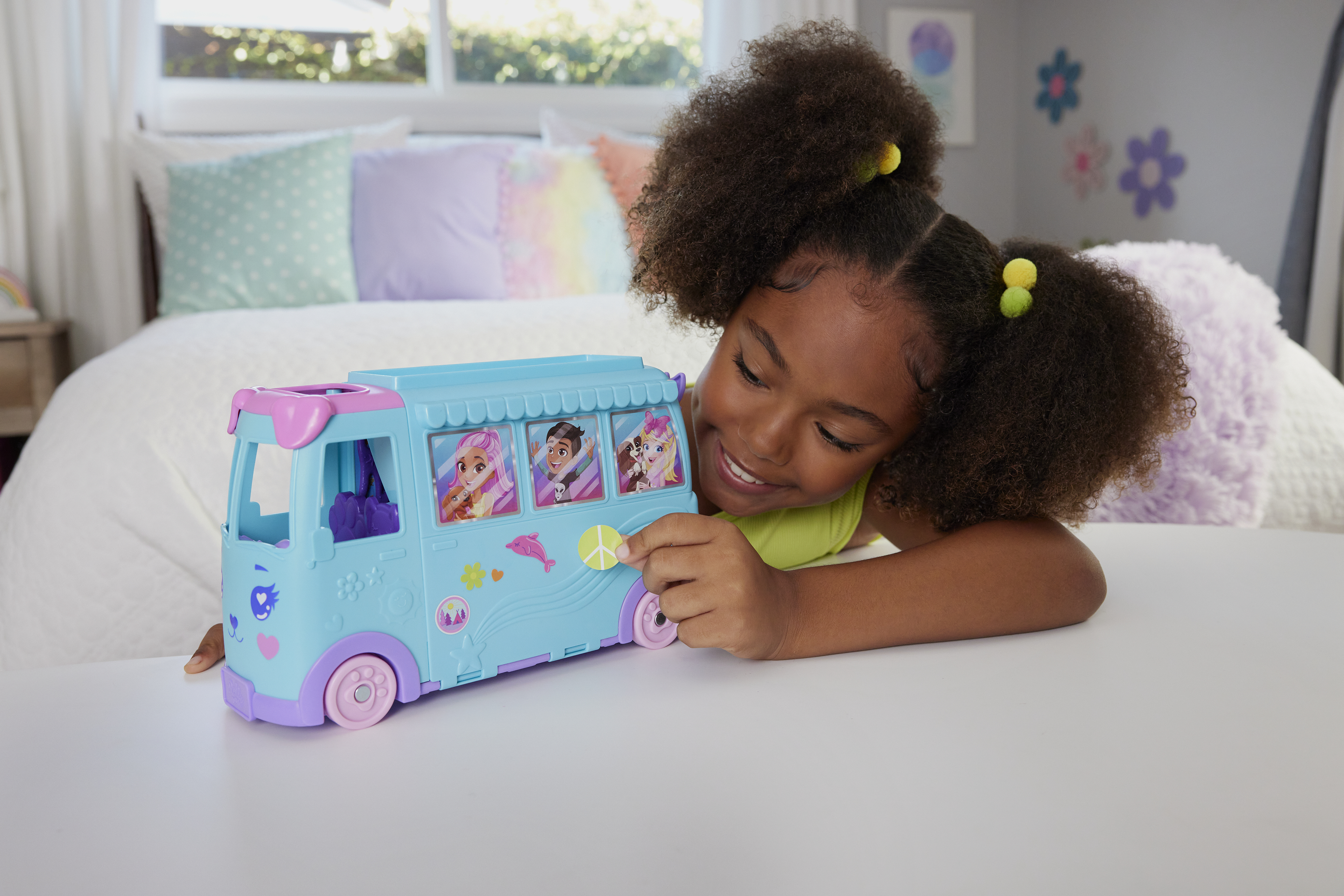 Dziewczyna uśmiecha się do niebieskiego autobusu-zabawki. Autobus ma różowe akcenty i zdjęcia ludzi w oknach. W tle widać łóżko.