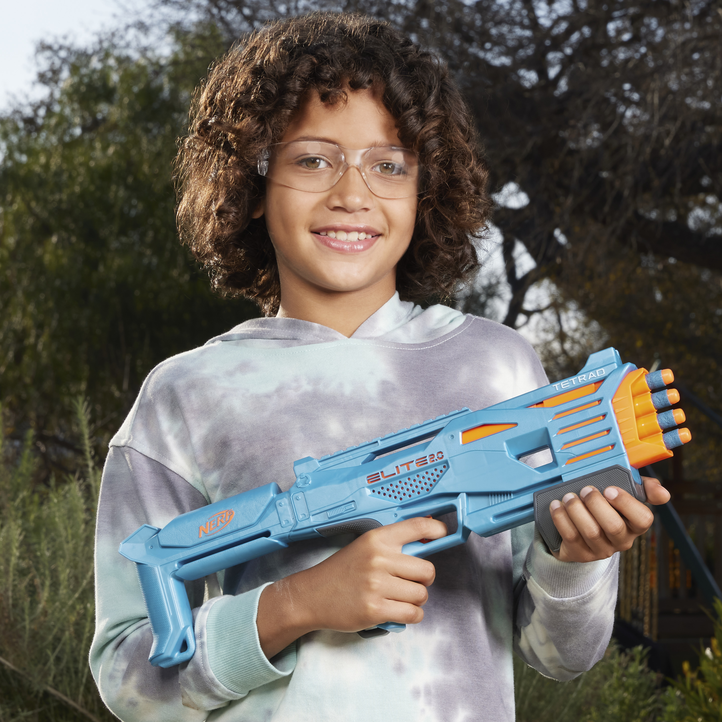 Un bambino sorridente tiene in mano una pistola Nerf blu. Lo sfondo include alberi e fogliame verde.