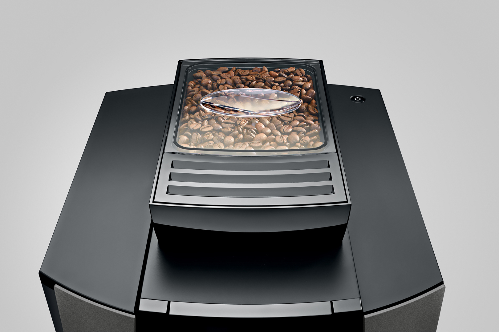 Eine schwarze Kaffeemaschine mit einem Bohnenbehälter gefüllt mit gerösteten Kaffeebohnen, auf grauem Hintergrund.