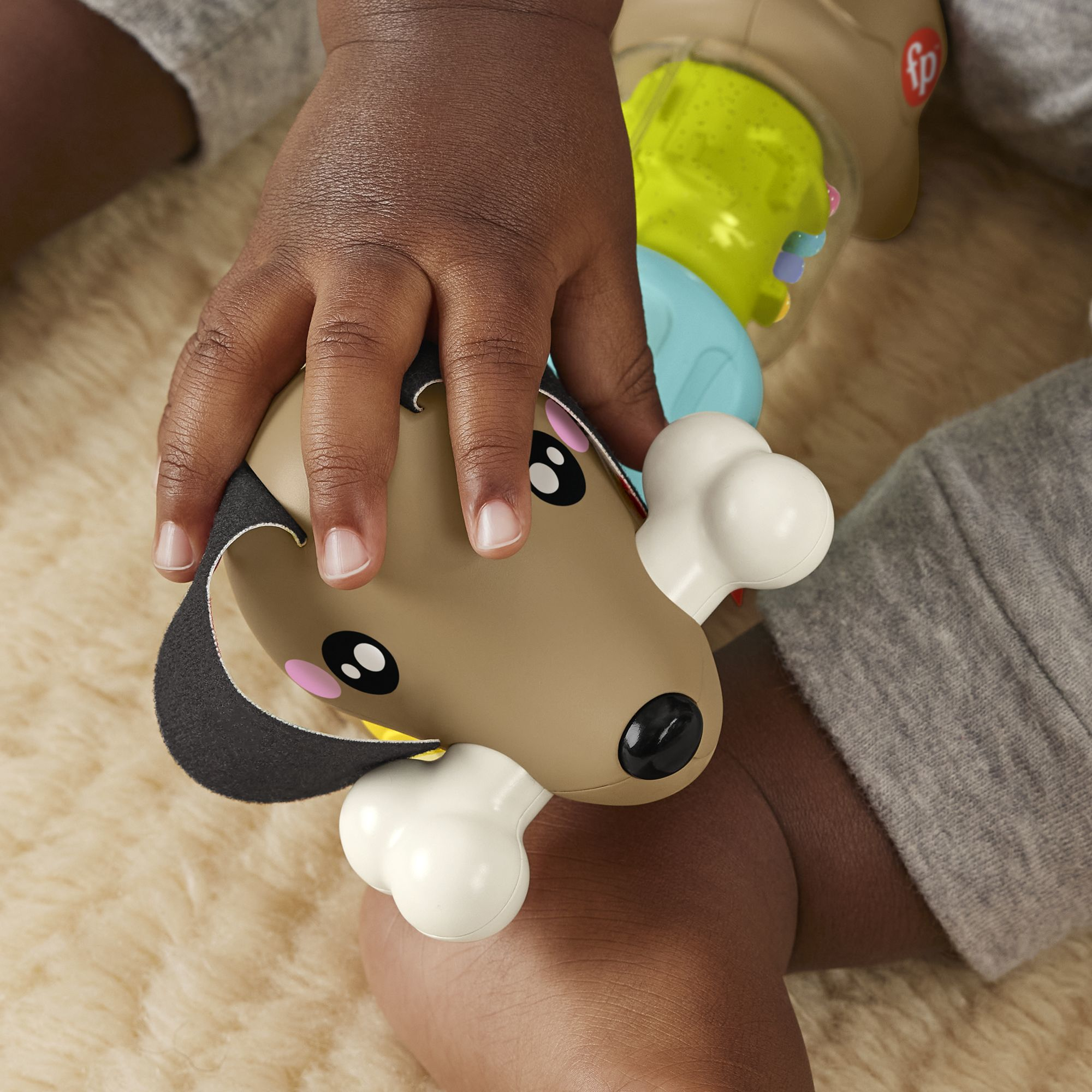 Kinderhände halten einen Spielzeughund mit Knochen, der Hund ist braun und hat schwarze Augen. Der Hintergrund ist eine beige Oberfläche und graue Kleidung.