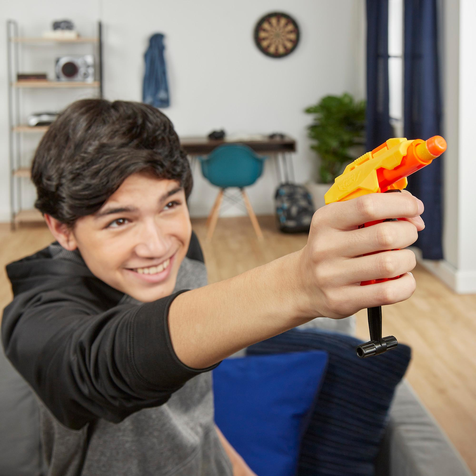 Ein lächelnder Teenager hält eine gelb-orange Spielzeugpistole und zielt damit nach vorne. Im Hintergrund sind eine Dartscheibe und Dekoration zu sehen.
