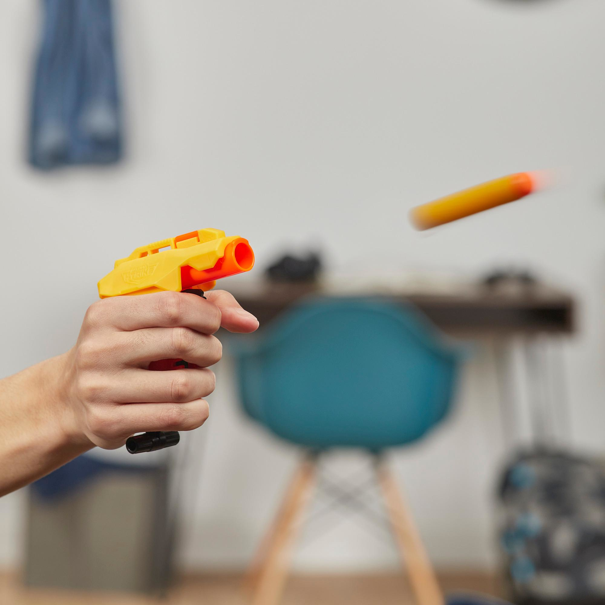 Eine Hand hält eine gelb-orangefarbene Spielzeugpistole, die einen orangefarbenen Pfeil abfeuert. Ein blauer Stuhl und verschwommener Hintergrund sind sichtbar.