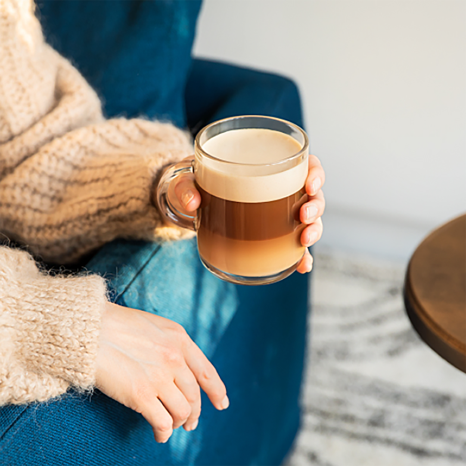 Osoba trzyma szklany kubek z warstwowym napojem kawowym. Ma na sobie beżowy sweter i niebieskie dżinsy. W pobliżu znajduje się drewniany stół.