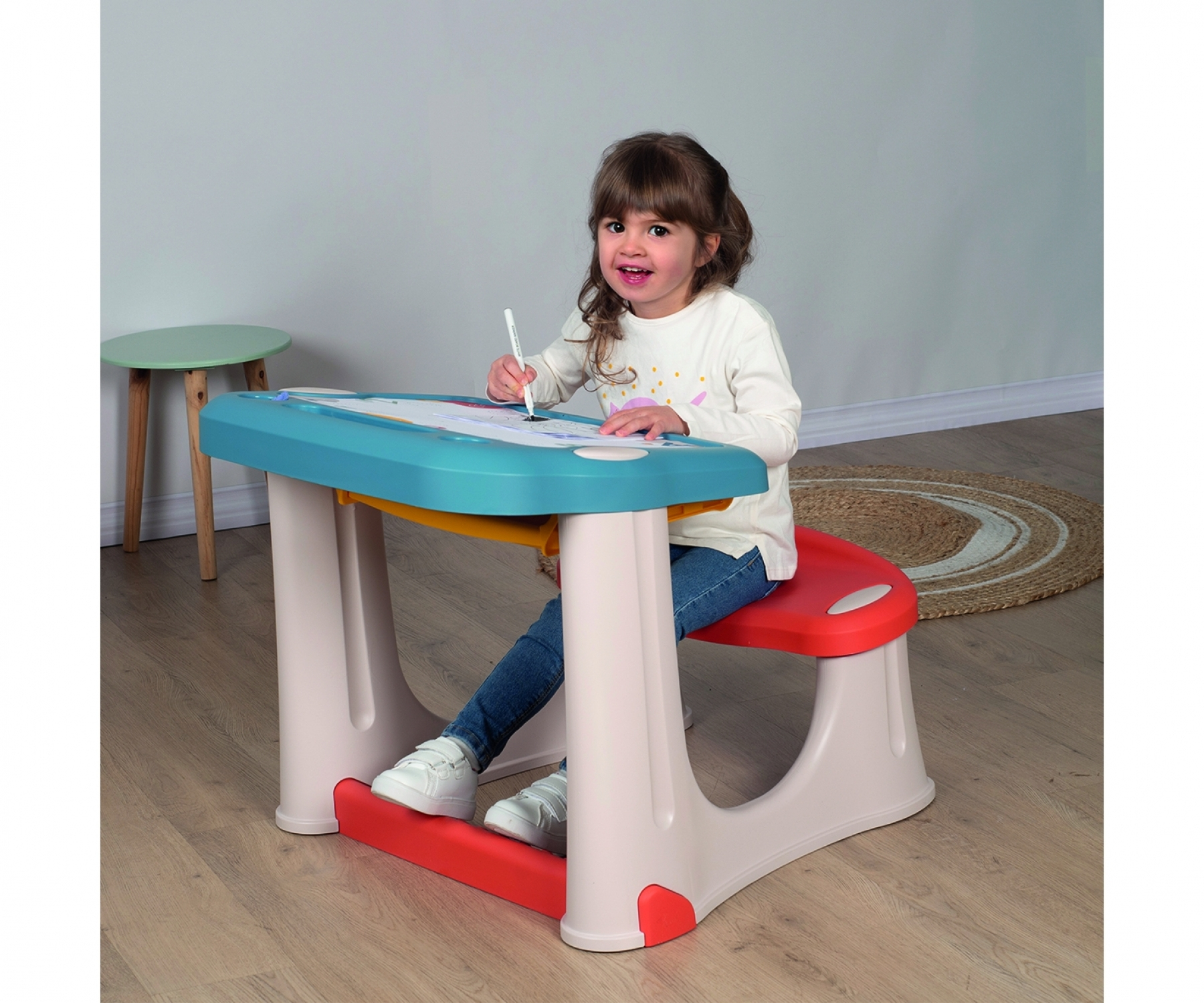 Młoda dziewczyna siedzi przy kolorowym biurku, rysując. Ma na sobie dżinsy i białą koszulę. W pobliżu znajduje się mały zielony stolik i dywan.