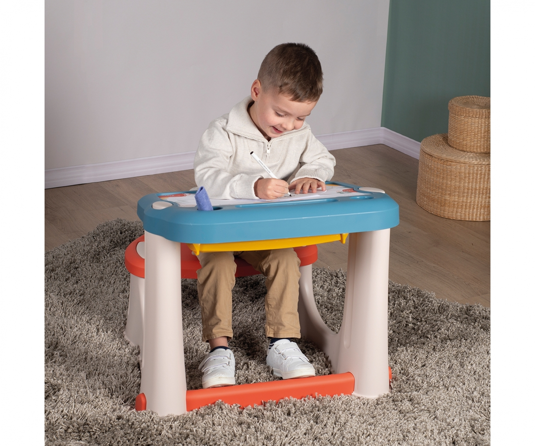 Mały chłopiec siedzi przy kolorowym biurku i rysuje. Biurko jest niebiesko-żółte. Ma na sobie biały sweter i brązowe spodnie.